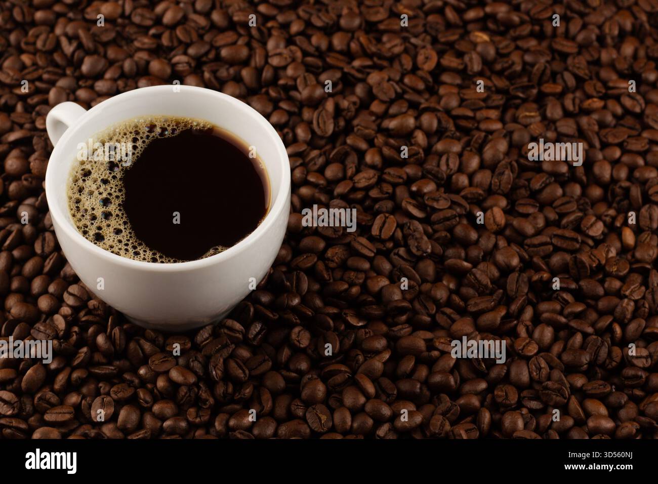 Eine weiße Tasse mit dunklem Kaffee steht auf einer Oberfläche, die mit aromatischen gerösteten Kaffeebohnen bedeckt ist und eine einladende Atmosphäre für Kaffee schafft Stockfoto