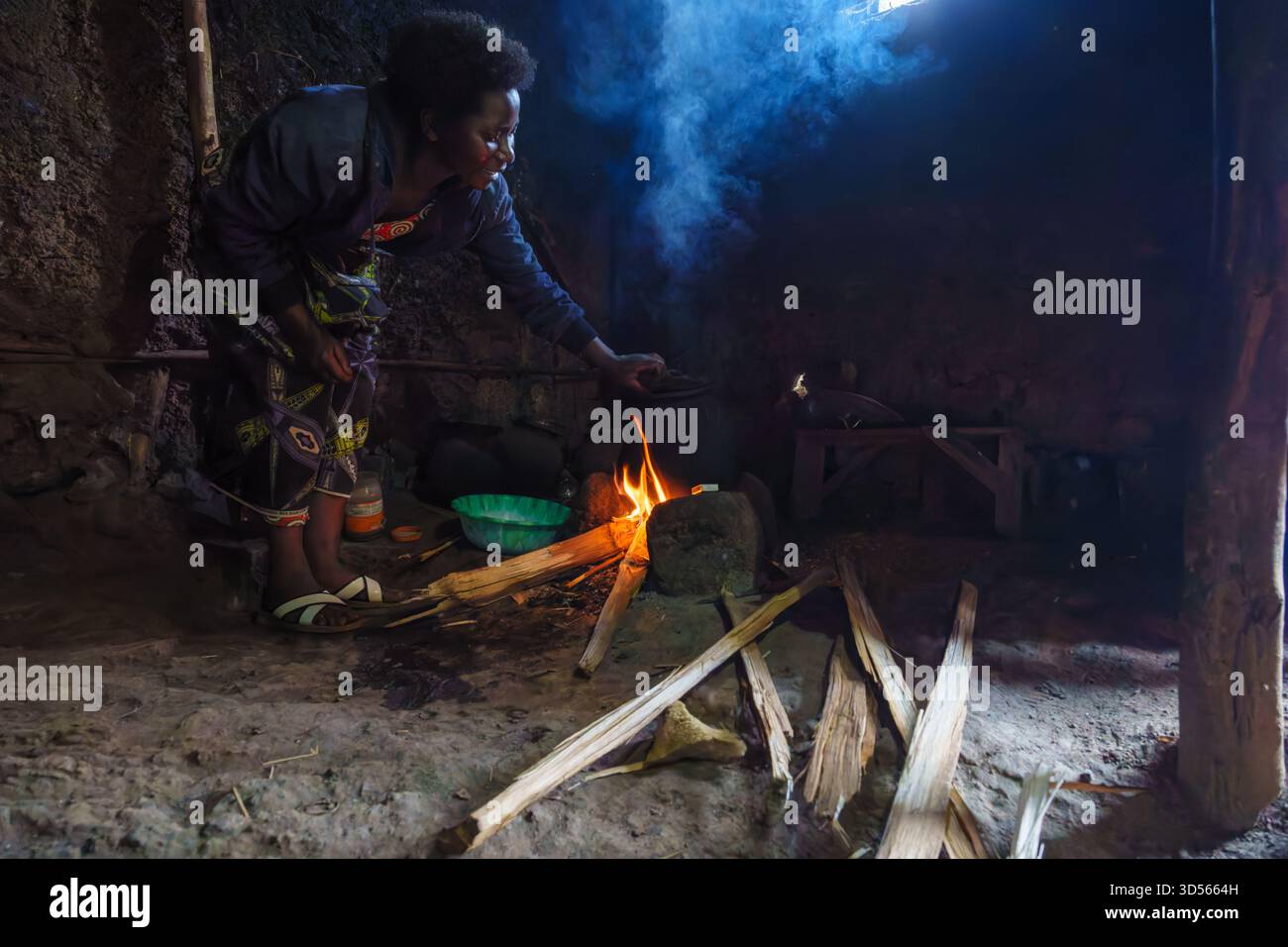 Gasura Ruanda - 8. September 2025; authentische Frau, die sich über rauchiges Feuer gebeugt hat und Essen aus der grünen Schüssel zum Feuer bringt, um auf traditionelle Weise ins Kochen zu kochen Stockfoto