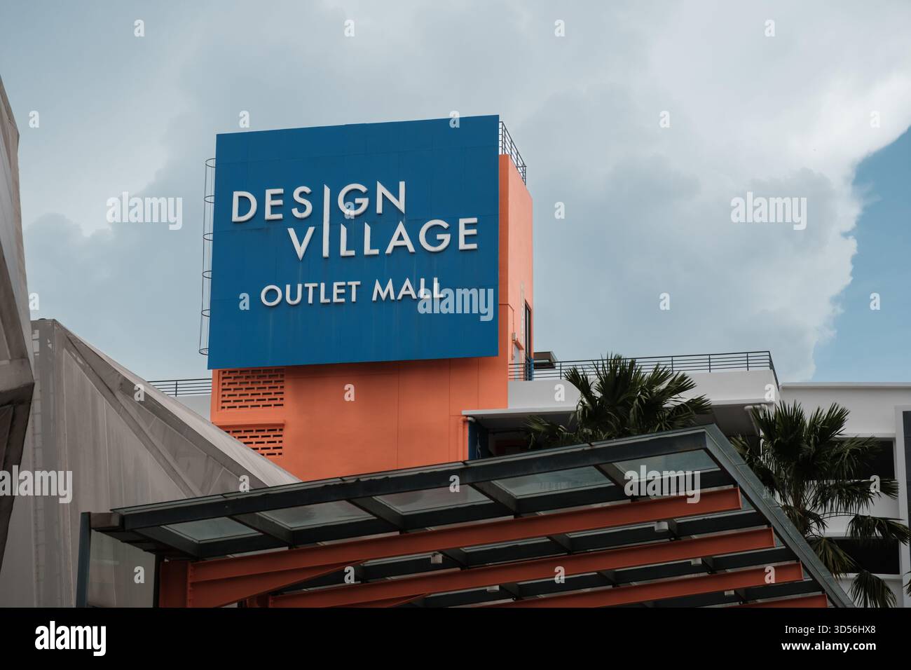 Penang, Malaysia - 16. Oktober 2025: Das Design Village Outlet Mall zeigt ein großes blau-orangefarbenes Schild. Wolken reflektieren Licht auf der Oberfläche Stockfoto