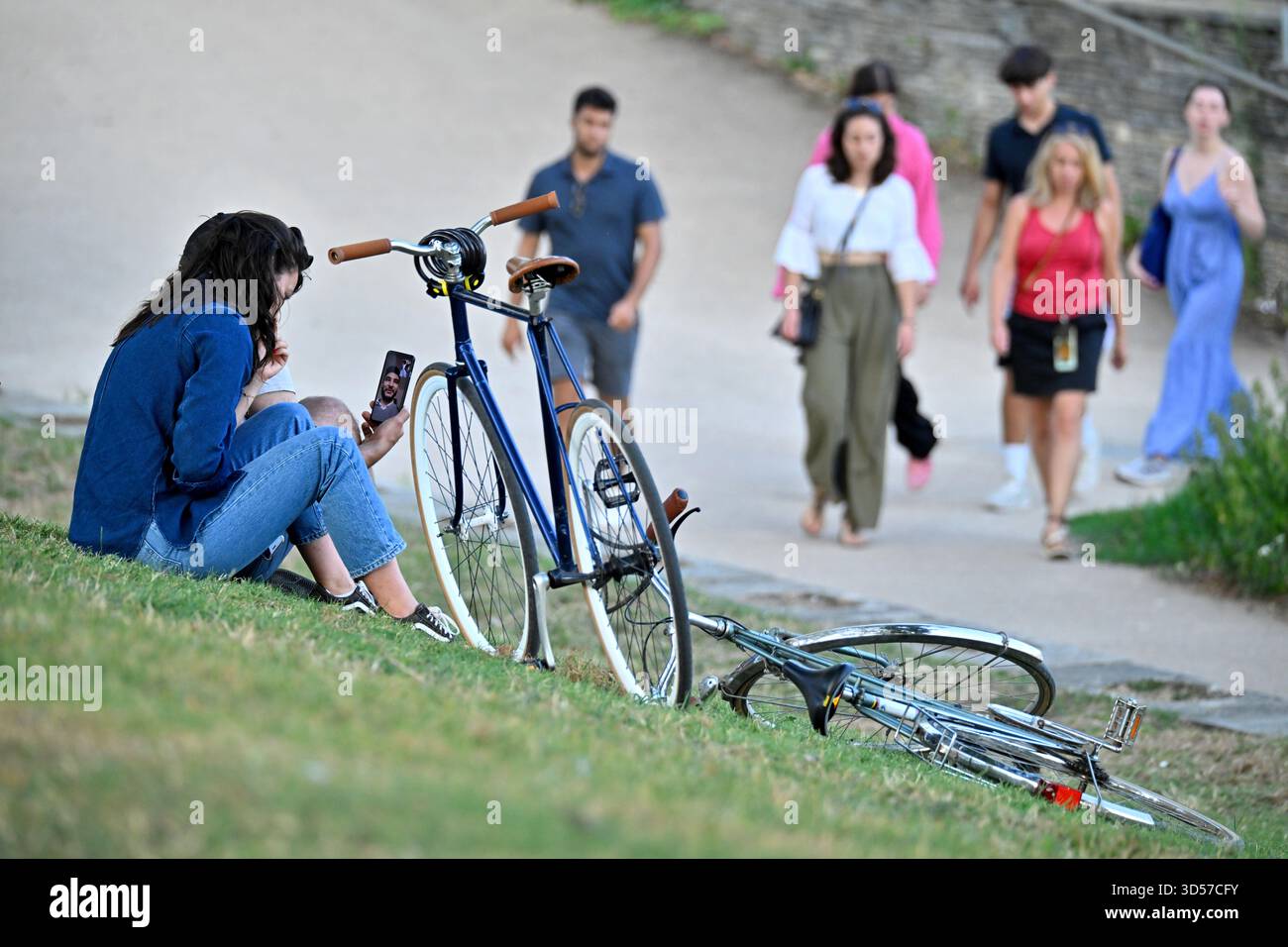 Angers (Nordwestfrankreich): Junge Radfahrerin, die auf einem abfallenden Rasen neben ihrem Fahrrad sitzt und per Videoanruf mit ihrem Freund über ein Smartphone spricht. G Stockfoto