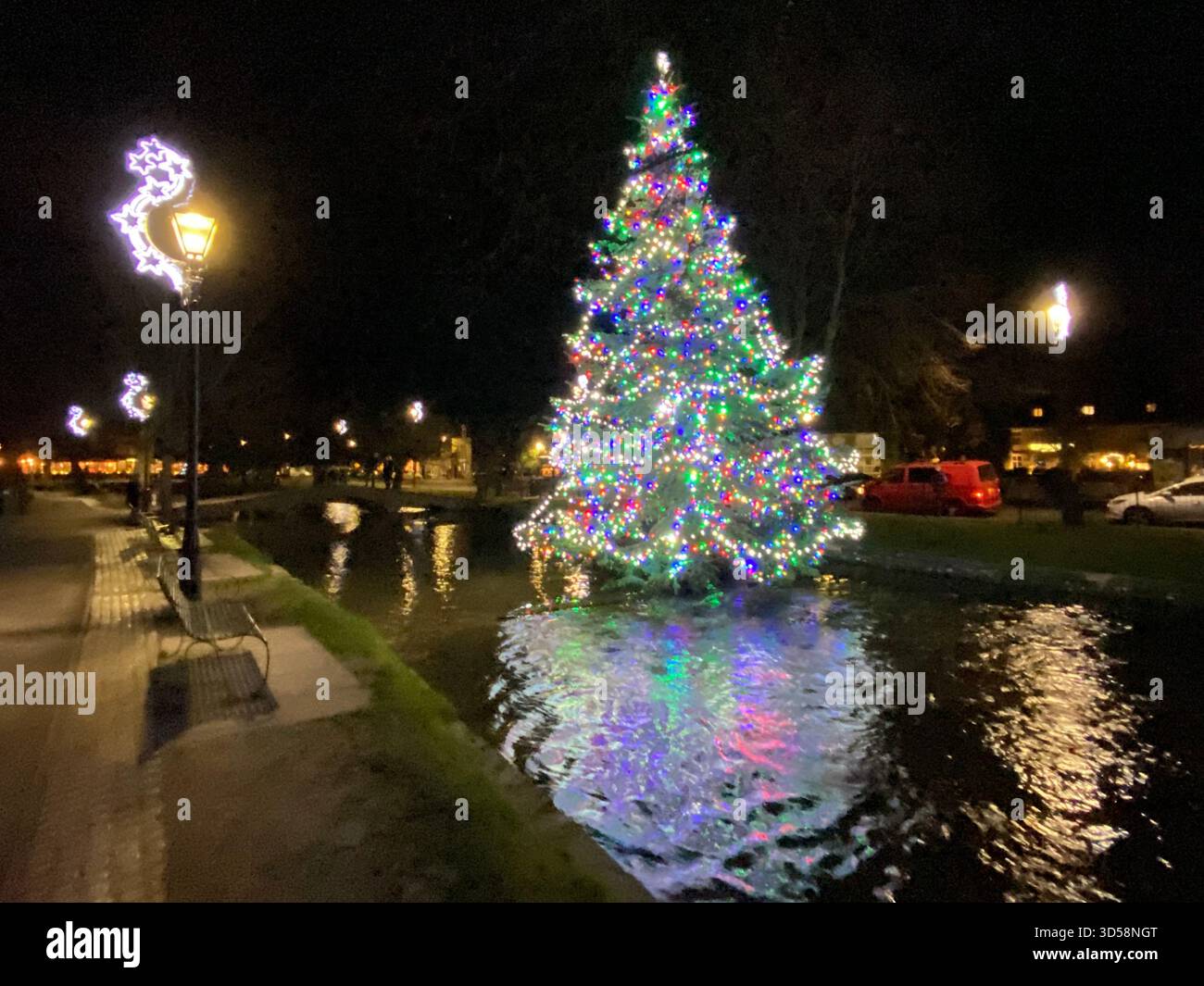 Ein Weihnachtsbaum in Bouton-on-the-Water steht inmitten des flachen Flusses Windrush, der wegen seiner „Venedig der Cotswolds“ genannt wird - Smartphone-aufgenommenes Stockfoto