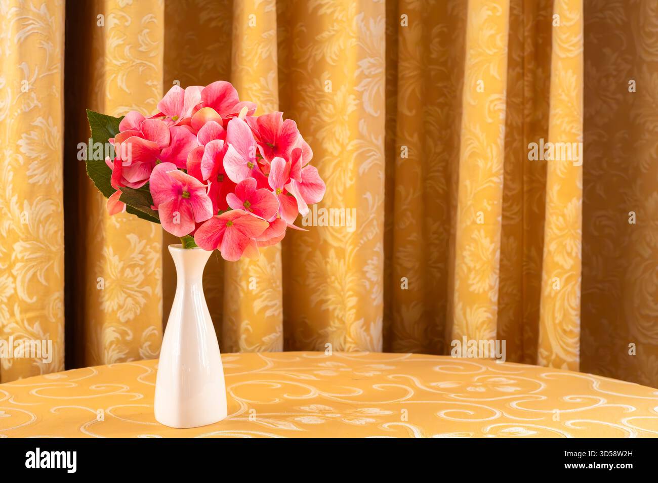 Eine weiße Vase mit rosa Blumen auf einer gemusterten gelben Tischdecke. Stockfoto
