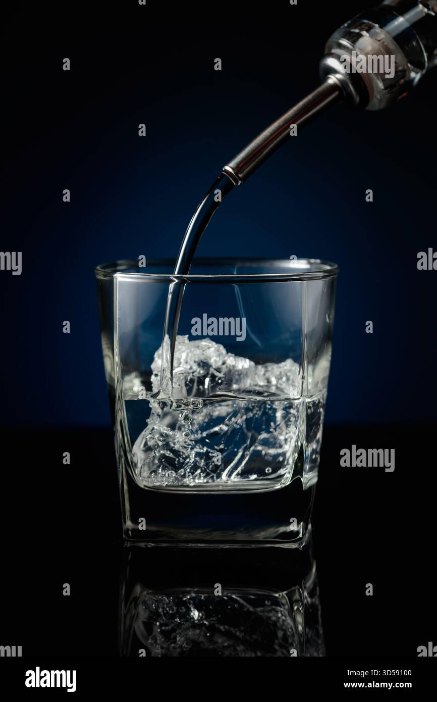 Kristallklarer Wodka oder Gin wird über Eis in ein Glas gegossen. Wodka mit Eis auf schwarzem reflektierendem Hintergrund. Stockfoto