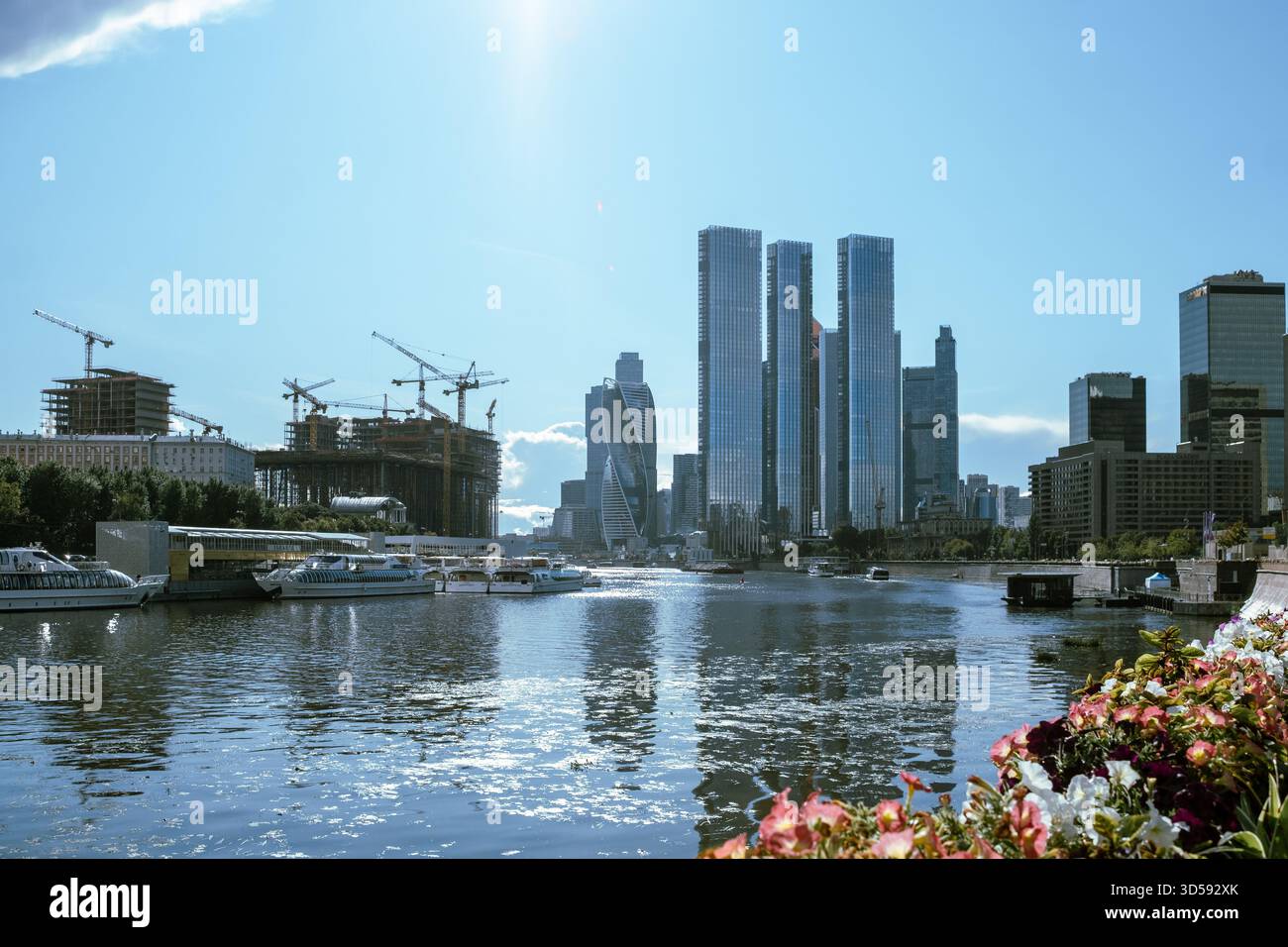 25. August 2025, Moskau, Russland. Wolkenkratzer erheben sich über einem ruhigen Fluss, der das Sonnenlicht reflektiert, während die Boote durch das Wasser fahren, umgeben von Konst Stockfoto