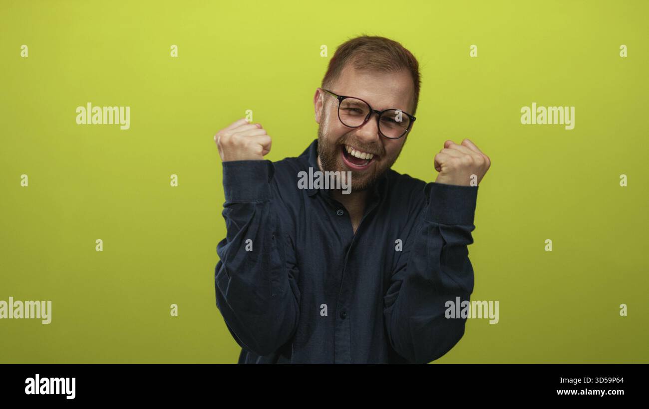 Mann mit geballten Fäusten und lachender Brille, in einem hellgrünen Atelier in einem marineblauen Hemd; Glücksfeier Erfolg. Stockfoto