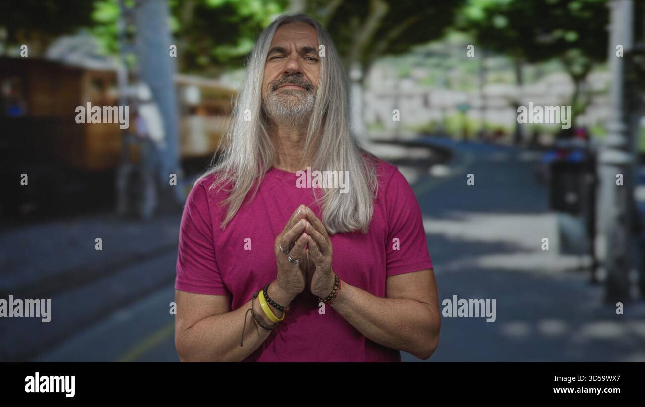 Mann mit gefalteten Händen und langen grauen Haaren, der auf einer sonnendurchfluteten Straße steht, Finger in der Nähe der Brust, rosafarbenes Hemd, Gelassenheit reflektiert. Stockfoto