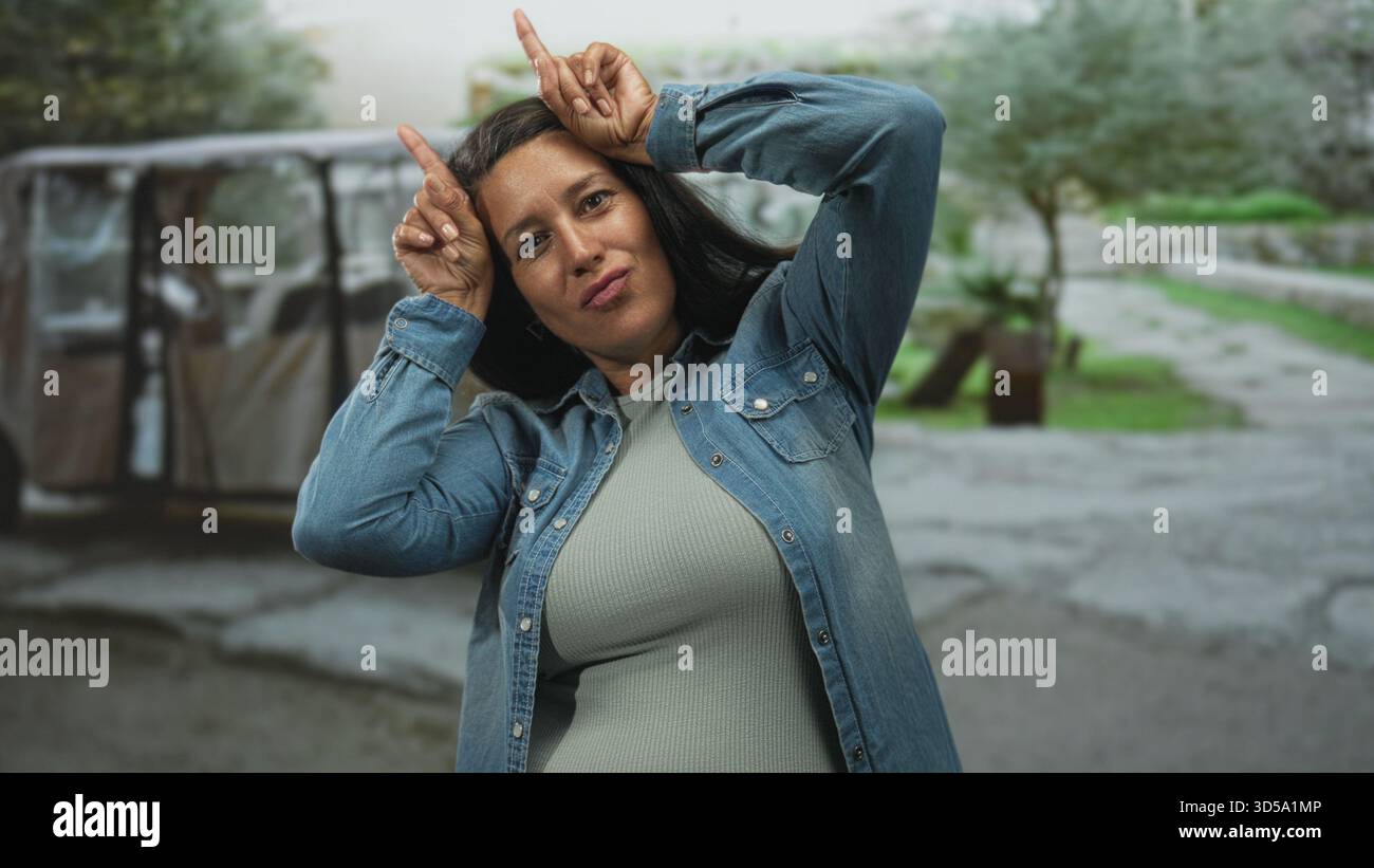 Frau, die Horngesten mit Zeigefingern an den Schläfen macht, Jeansjacke und geripptes Oberteil trägt, auf einer zerrissenen Straße in der Nähe eines geparkten Karren und Baumes steht Stockfoto