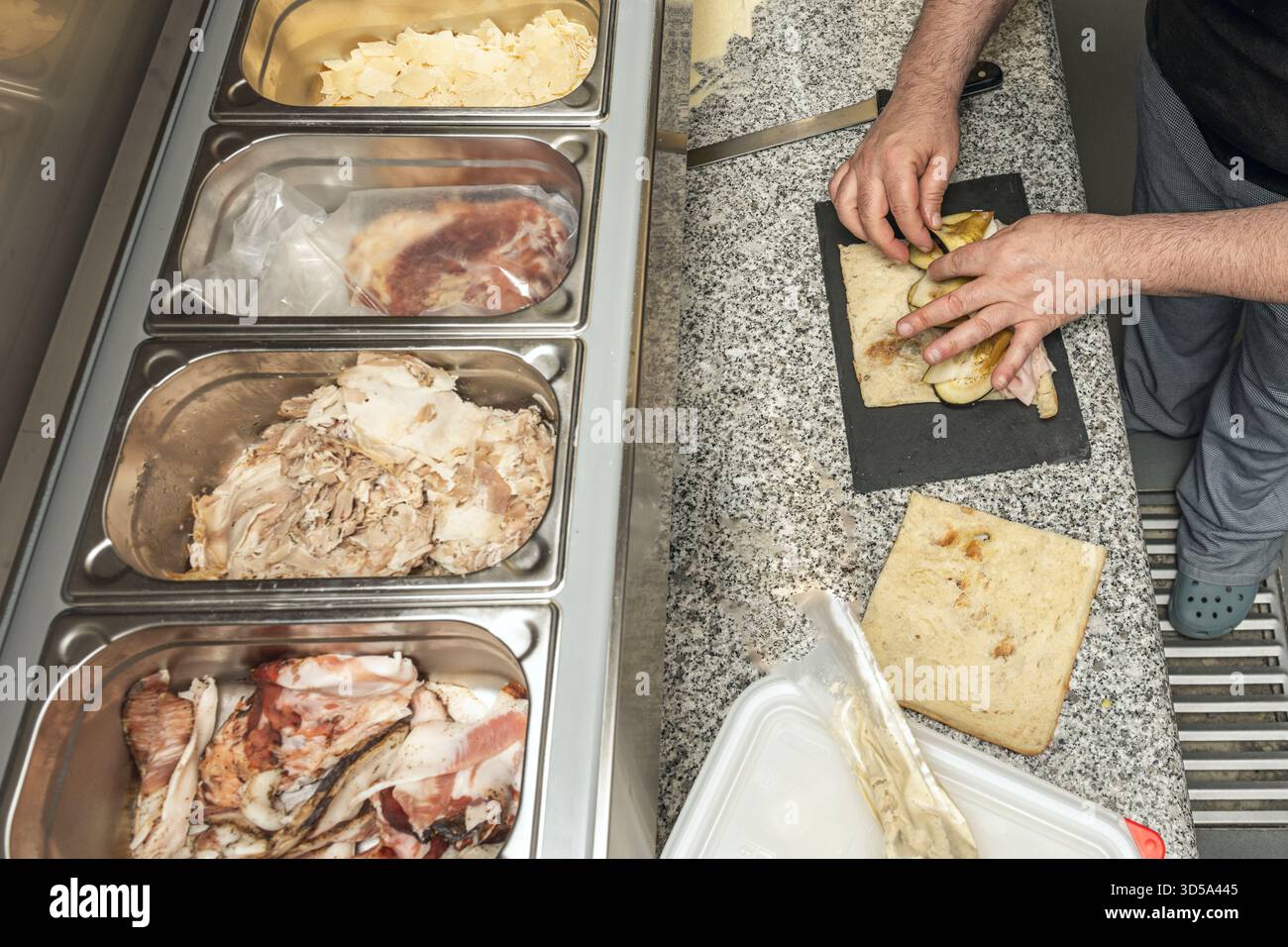 Die bemehlten Finger des Bäckers drücken die Kalamata-Oliven sanft auf die Oberfläche der Focaccia, wodurch ein harmonisches Muster auf der elastischen, backen-fertigen Oberfläche entsteht Stockfoto