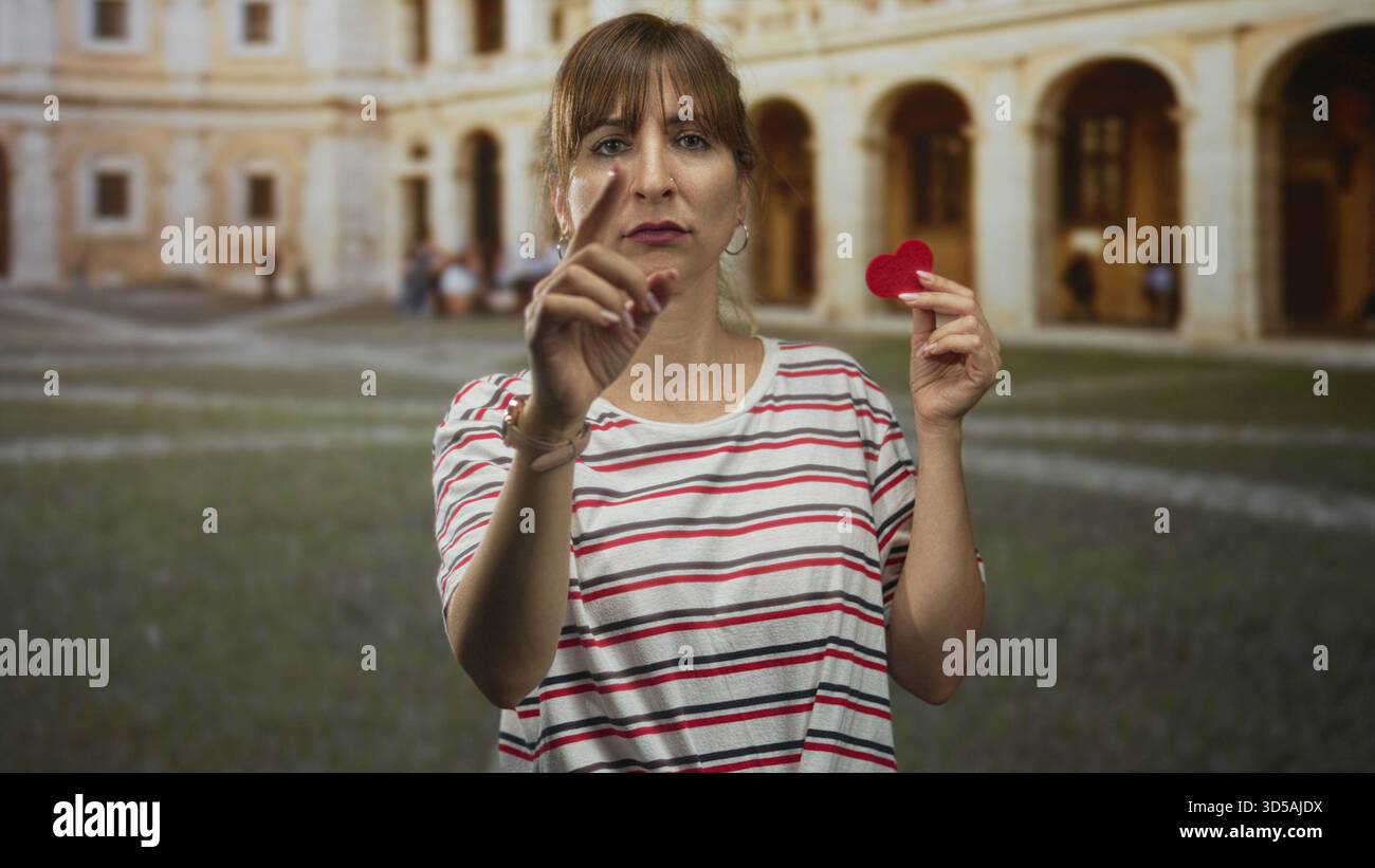 Die Frau zeigt den Finger nach vorne und hält ein kleines rotes Filzherz neben den Bögen des Innenhof des Altstadtgebäudes, trägt ein gestreiftes Hemd und Creolen; Liebe Stockfoto