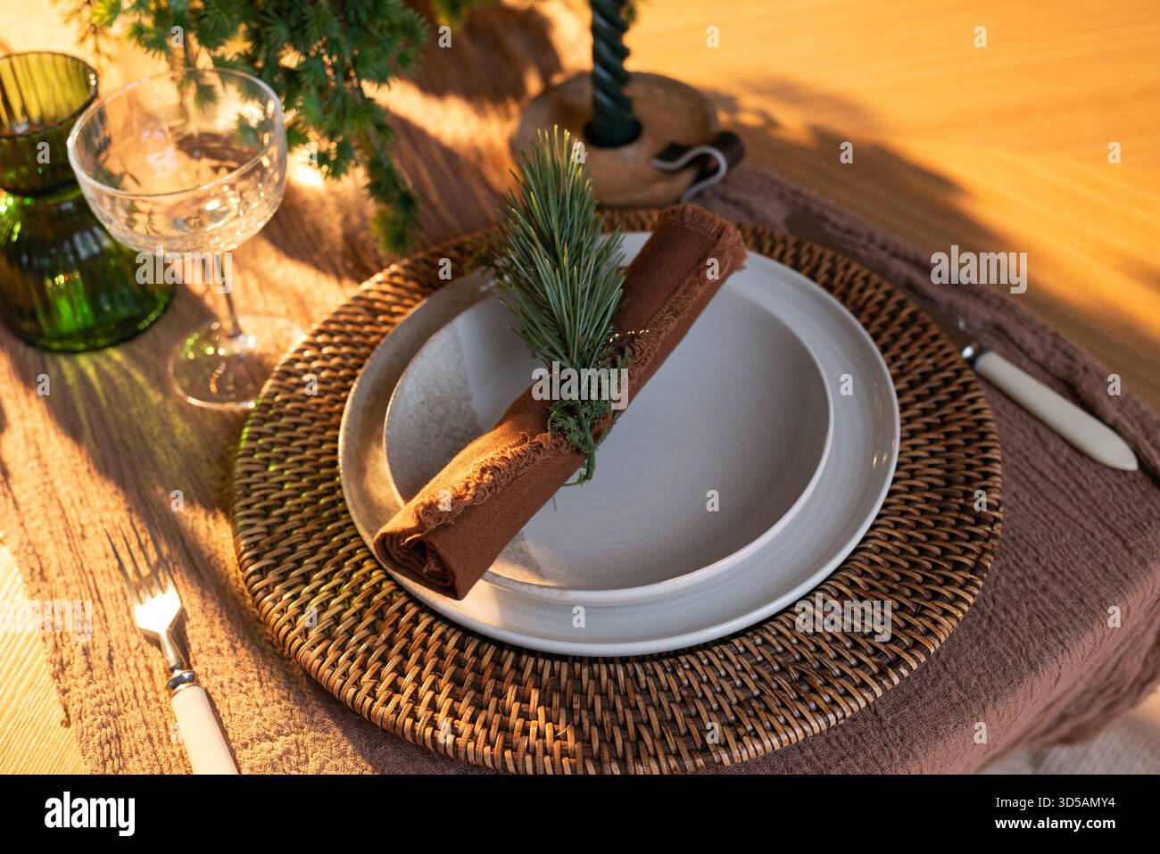 Festliche Tischdekoration mit gewebtem Ladegerät, Keramikplatten, rustikaler Serviette und grüner Dekoration in warmer Weihnachtsbeleuchtung Stockfoto
