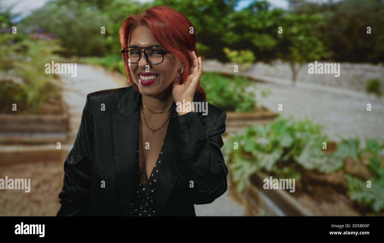 Frau von Hand zu Ohr hört und lächelt in einem Straßengarten mit Pflanzgefäßen und Grün; Freude Verbindung Wärme. Stockfoto