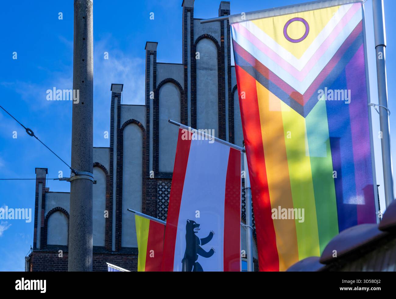 Die Progress Pride-Flagge neben der Berliner Landesflagge und anderen öffentlichen Bannern, Berlin-Friedrichshain, Deutschland. Pride Flag, Regenbogenflagge, Vielfalt. Stockfoto