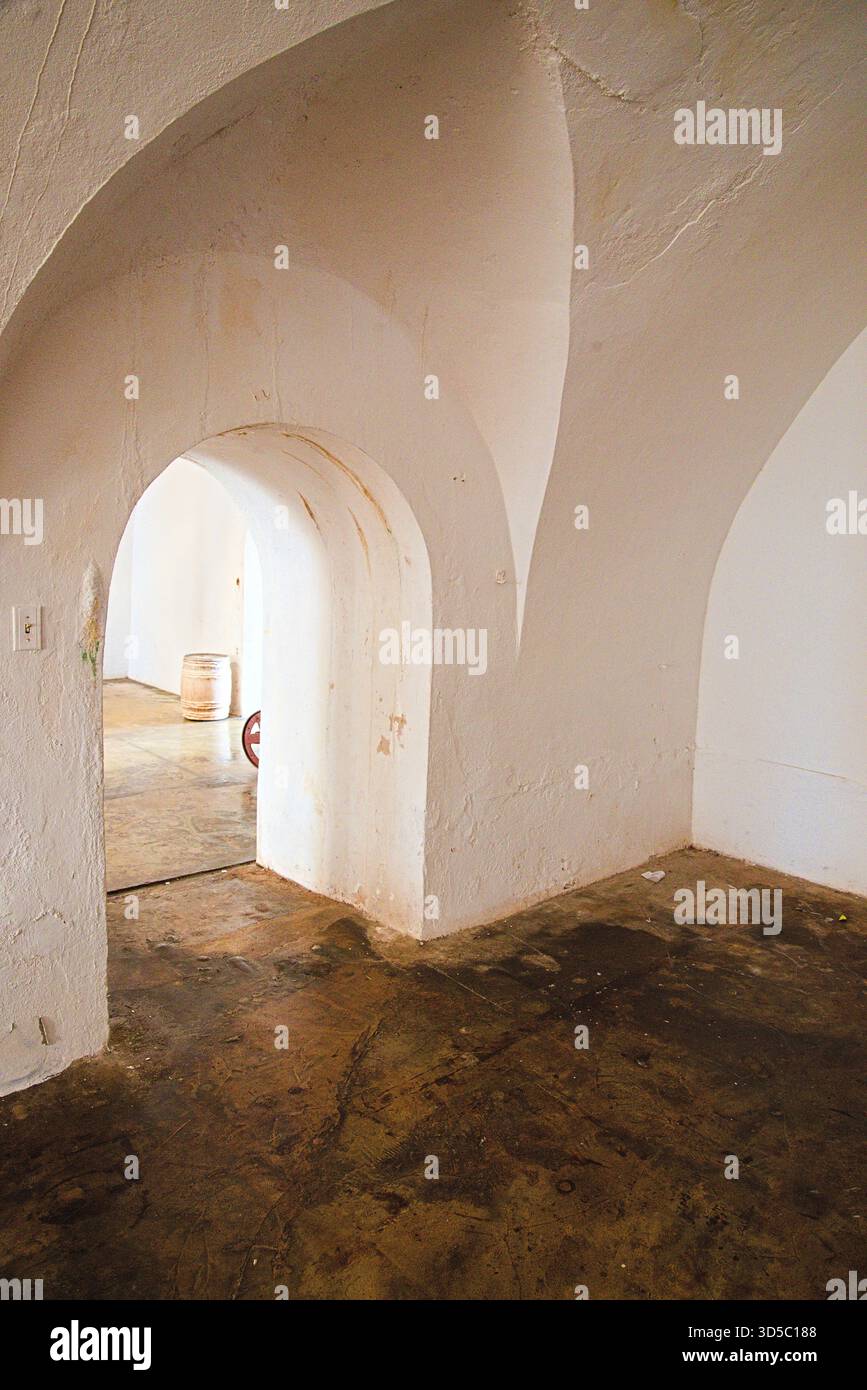 Bogenförmige Eingänge führen von einem schwach beleuchteten Raum in einen helleren Raum, der die rustikale Architektur unterstreicht. Der Boden zeigt ein verwittertes Aussehen. Stockfoto