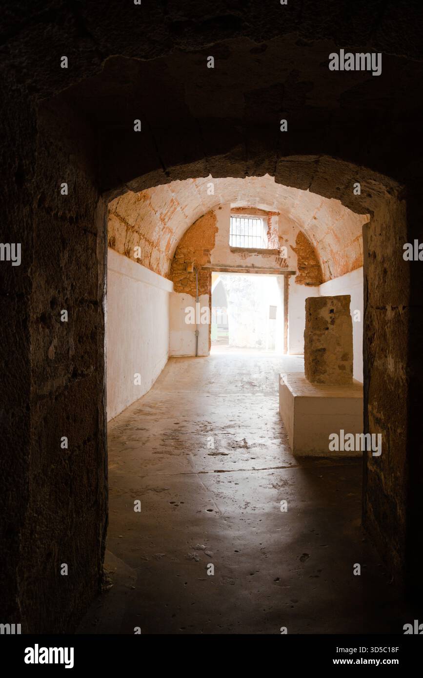 Ein schwach beleuchteter Steinkorridor führt zu einem hellen Außenbereich mit alten Ziegelbögen und rustikalen Wänden, die historischen Charakter verleihen. Stockfoto