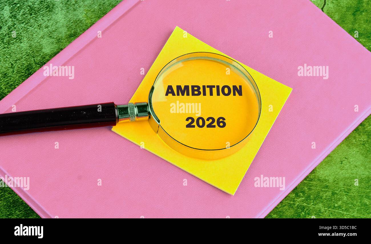 Eine Lupe konzentriert sich auf die Worte Ambition 2026, die auf einer gelben Haftnotiz geschrieben sind. Die Notiz befindet sich auf einem rosafarbenen Buch, das auf einer grünen strukturierten Brandung steht Stockfoto