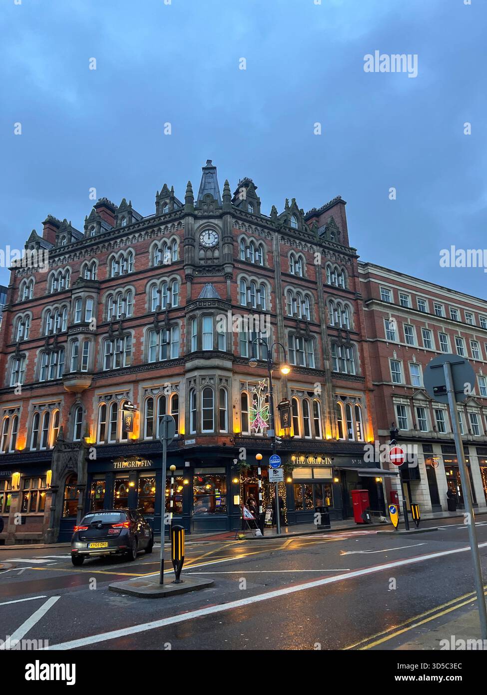 Abendliche Straßenszene mit einem historischen Eckgebäude in Leeds, England, mit Geschäften und Verkehr. Städtische Architektur und Stadtleben bei nassem Wetter. Stockfoto