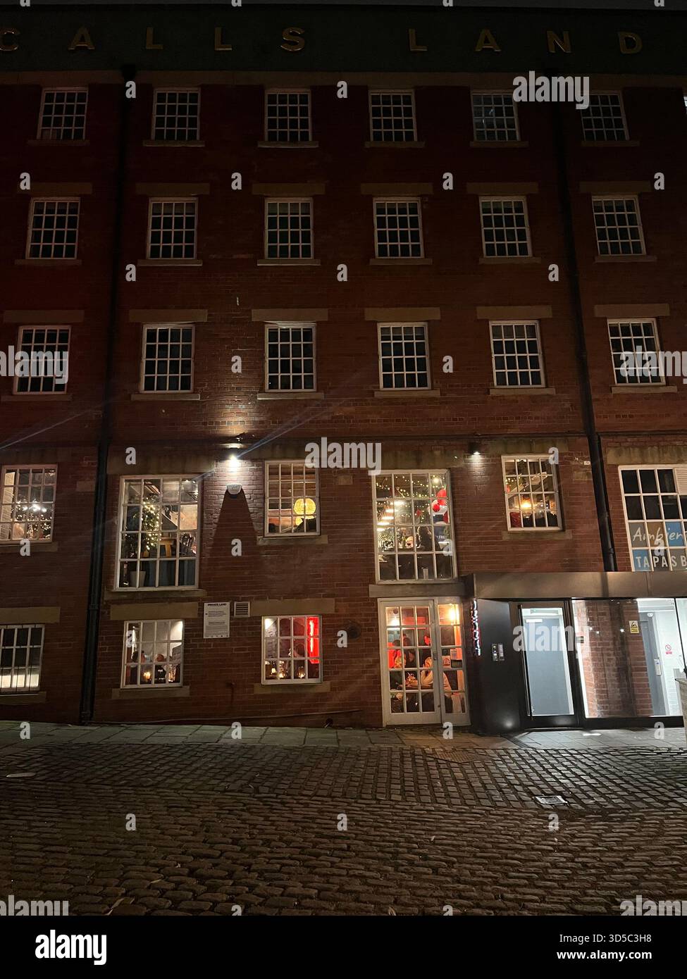 Historisches rotes Backsteingebäude mit beleuchteten Fenstern bei Nacht in Leeds, England. Atmosphärische urbane Szene mit Kopfsteinpflasterstraße und warmer Inneneinrichtung - Smartphone-aufgenommenes Stockfoto