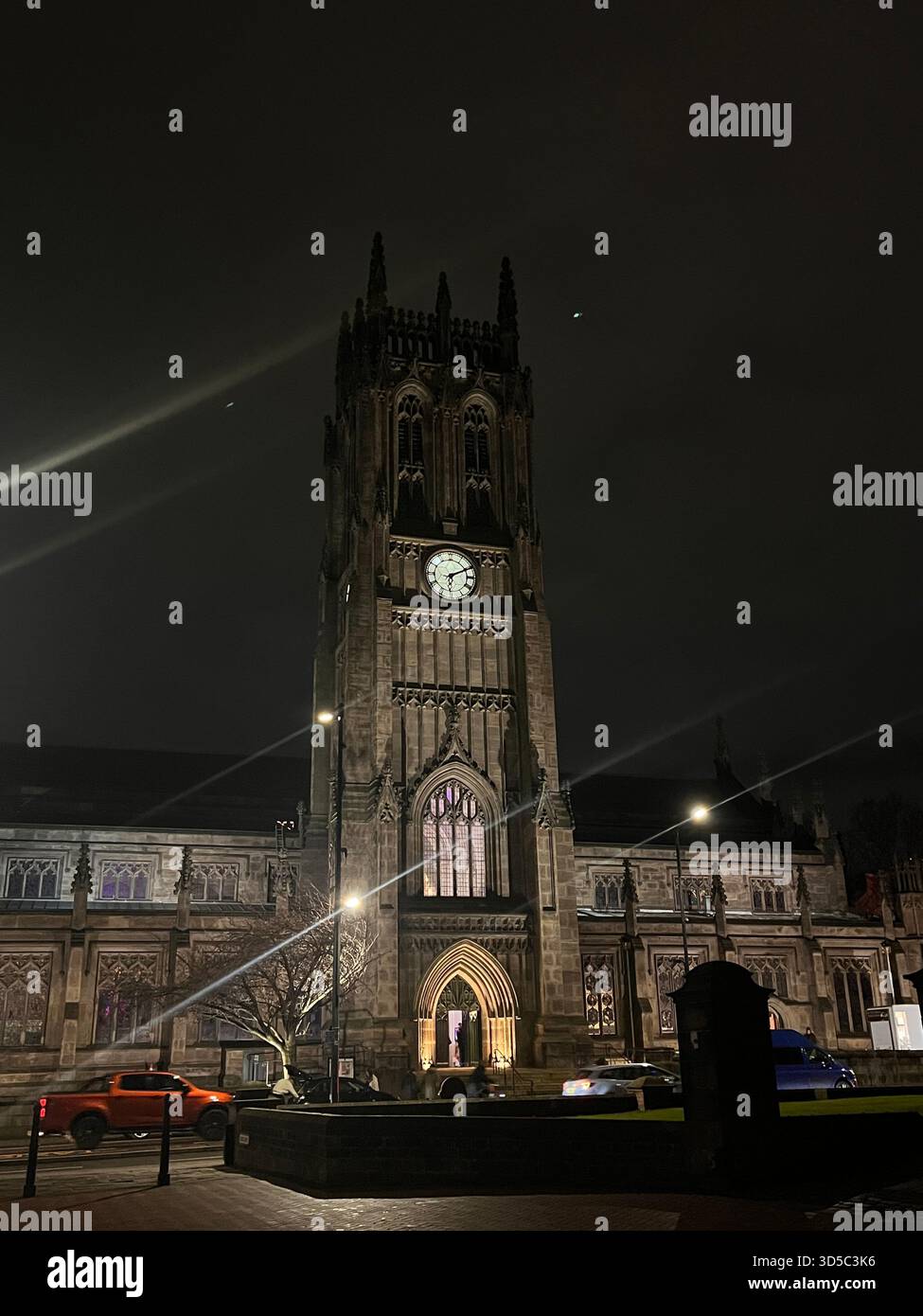 Hoher historischer Kirchturm mit beleuchtetem Zifferblatt, fotografiert bei Nacht in Leeds, England, umgeben von dunkler urbaner Landschaft. - Smartphone-aufgenommenes Stockfoto