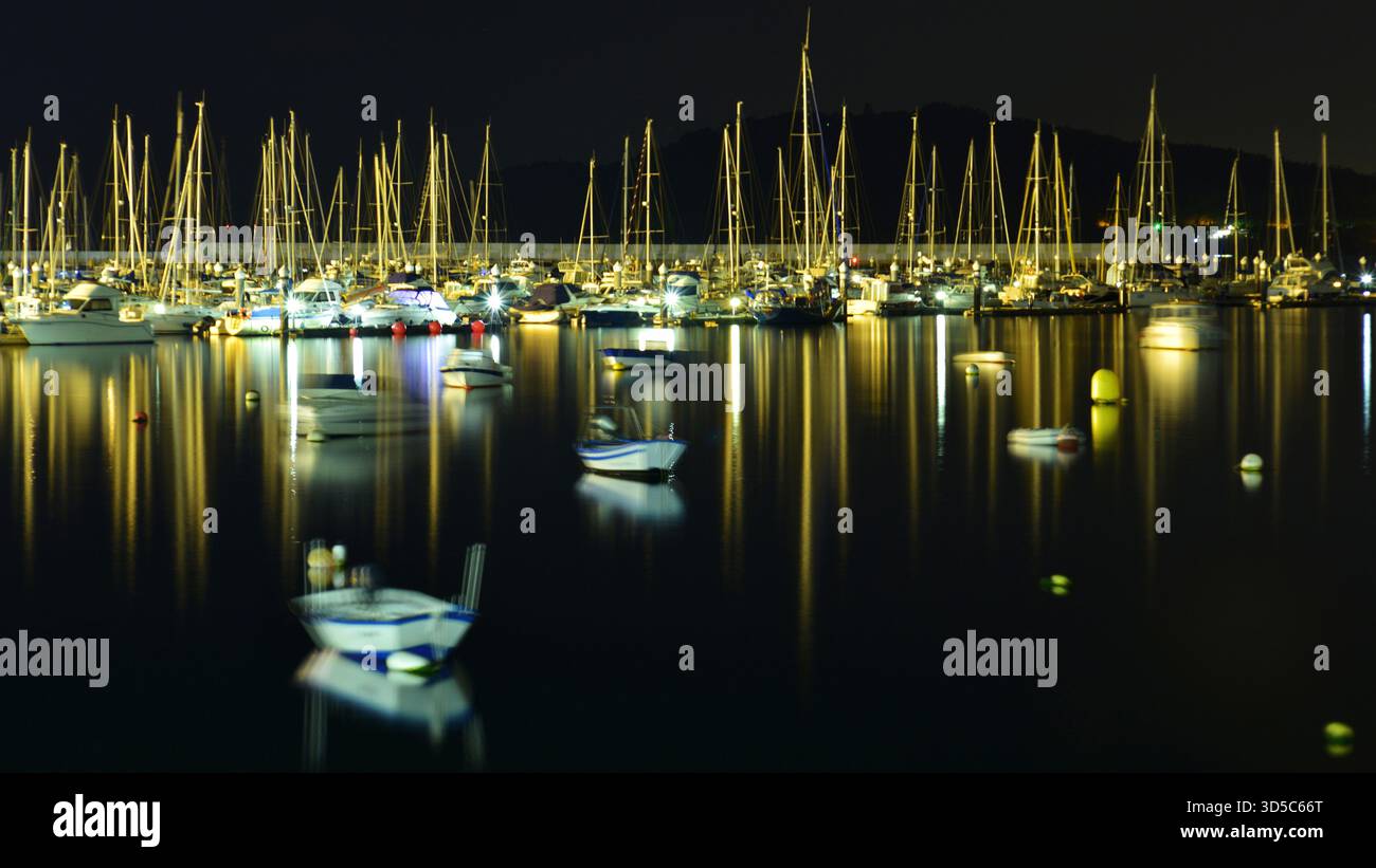 Nachtszene im Hafen von Baiona. Segelboote und Masten spiegeln sich im Wasser. Ein Boot im Vordergrund, bewegt von sanften Wellen. Stockfoto