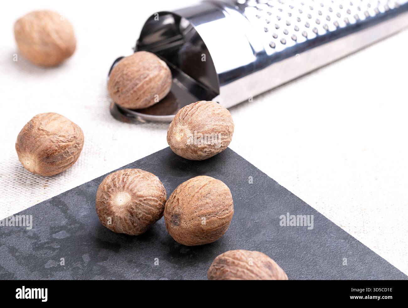 Ganze Muskatnüsse, auf einer Steinplatte, neben einer Muskatreibe, auf Leinen. Einige getrocknete Samen von duftenden oder echten Muskatnuss-Früchten, Myristica-Düfte. Stockfoto