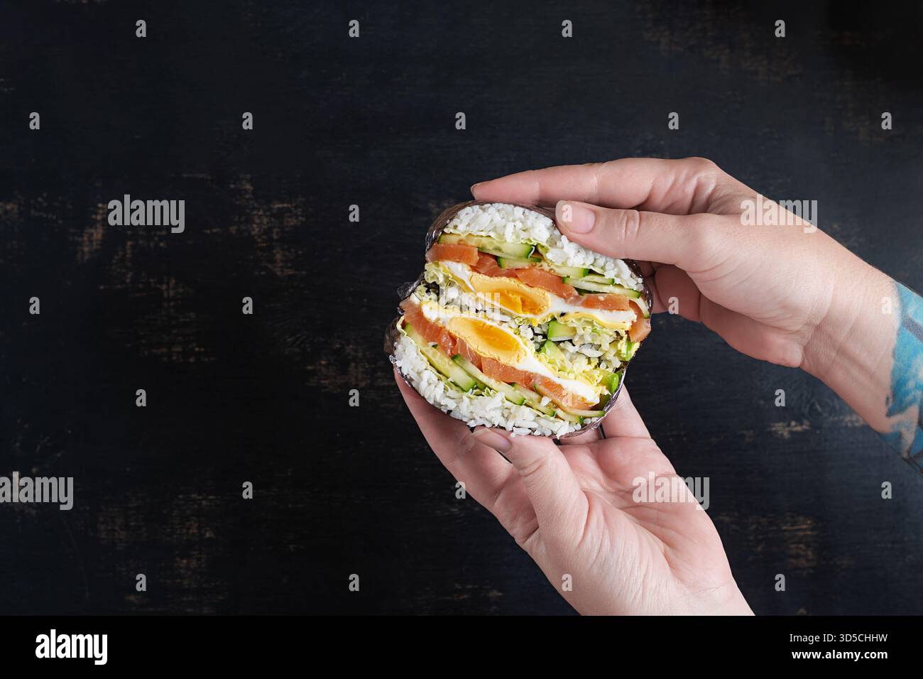 Ein kreativer Sushi-Burger mit Lachs, Spiegelei, Käse, Gurke und Avocado in den Händen einer Frau. Stockfoto