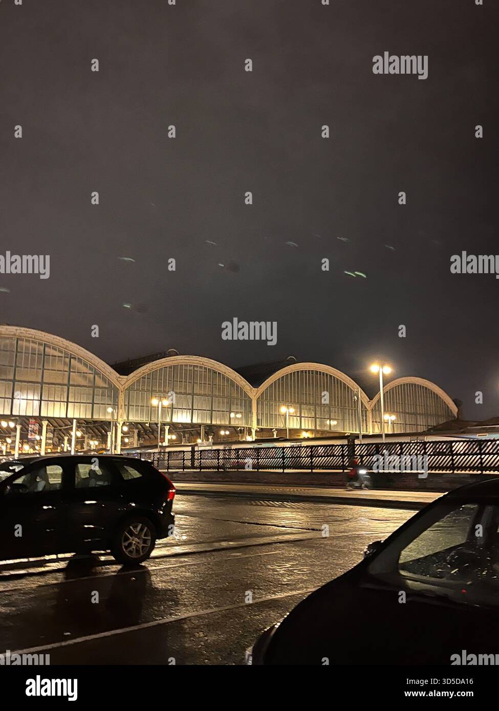 Nachtansicht auf ein modernes Gebäude mit geschwungenem Dach in der Nähe des Ufers in Hull, England, mit Reflexionen auf nassem Pflaster und geparktem Auto im Vordergrund. - Smartphone-aufgenommenes Stockfoto