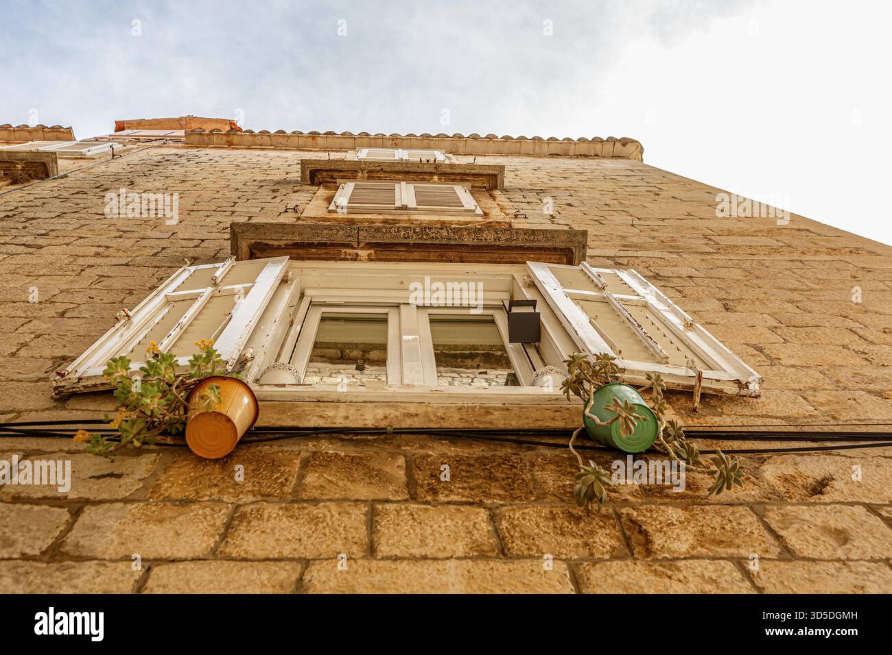 Blick nach oben auf ein mediterranes Steinhaus mit offenen weißen Fensterläden und kleinen Topfpflanzen unter dem Fenster. Die rustikale Architektur und das warme Licht spiegeln den Charme der südlichen Küstenstädte wider. Stockfoto