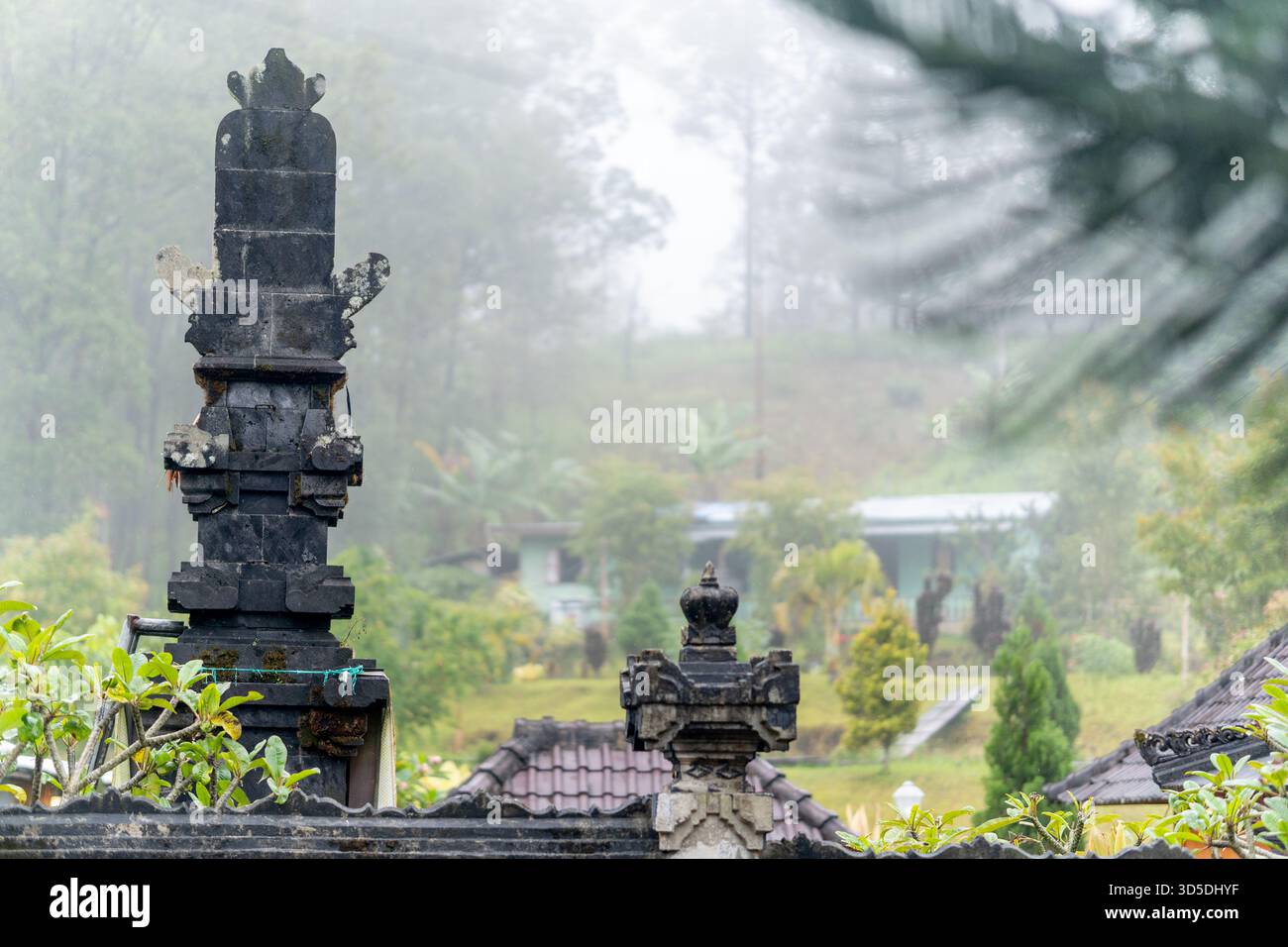 Nahaufnahme eines traditionellen balinesischen Tempels aus dunklem Stein mit Schnitzereien im Vordergrund mit einer nebeligen, grünen tropischen Hügellandschaft im Hintergrund. Stockfoto