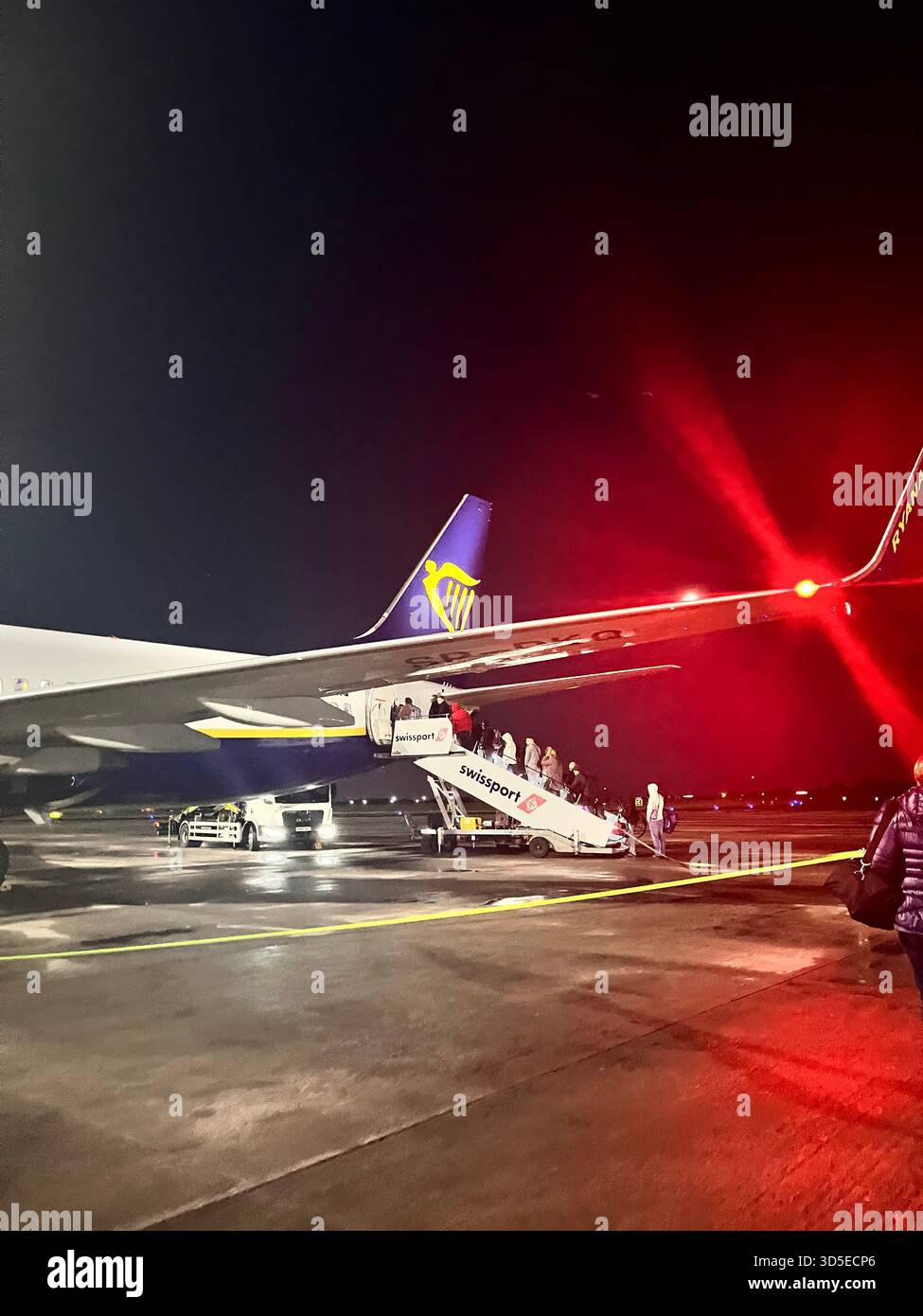 Dramatische nächtliche Szene eines Ryanair-Flugzeugs auf dem Asphalt in Leeds, beleuchtet von intensiven roten Lichtern und umgeben von Reflexionen auf dem nassen Asphalt. Stockfoto