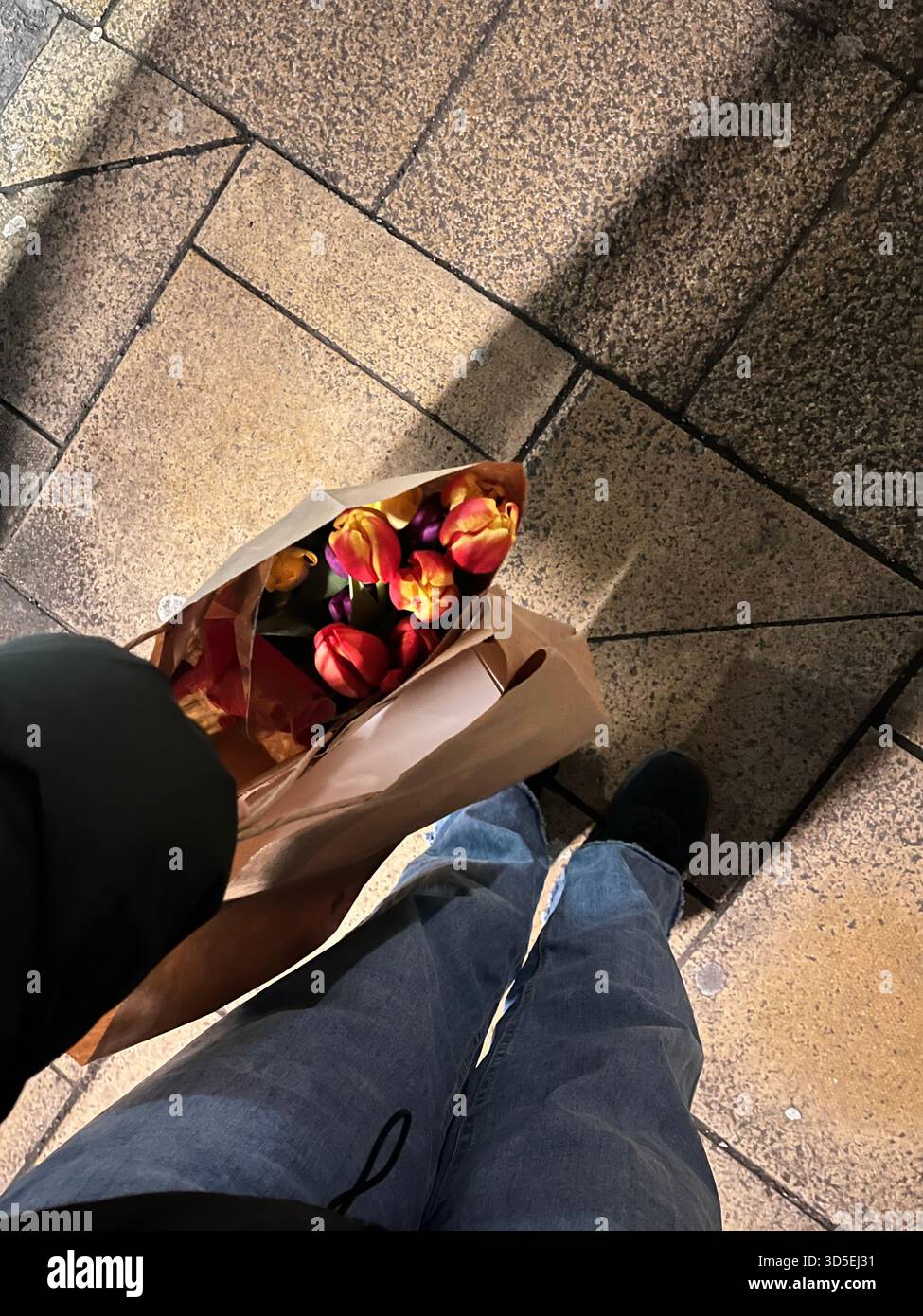 Eine Person, die auf einer gepflasterten Straße spaziert und Einkaufstaschen mit Tulpen hält. Die Szene zeigt einen alltäglichen urbanen Moment in Hull, England - Smartphone-aufgenommenes Stockfoto
