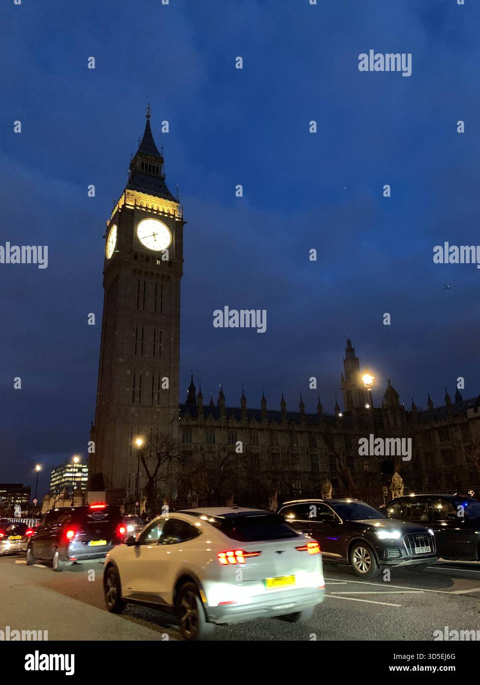 Ein hoher beleuchteter Uhrenturm steht vor dem dunklen Abendhimmel, umgeben von fahrenden Autos und hellen Lichtern der Stadt. - Smartphone-aufgenommenes Stockfoto