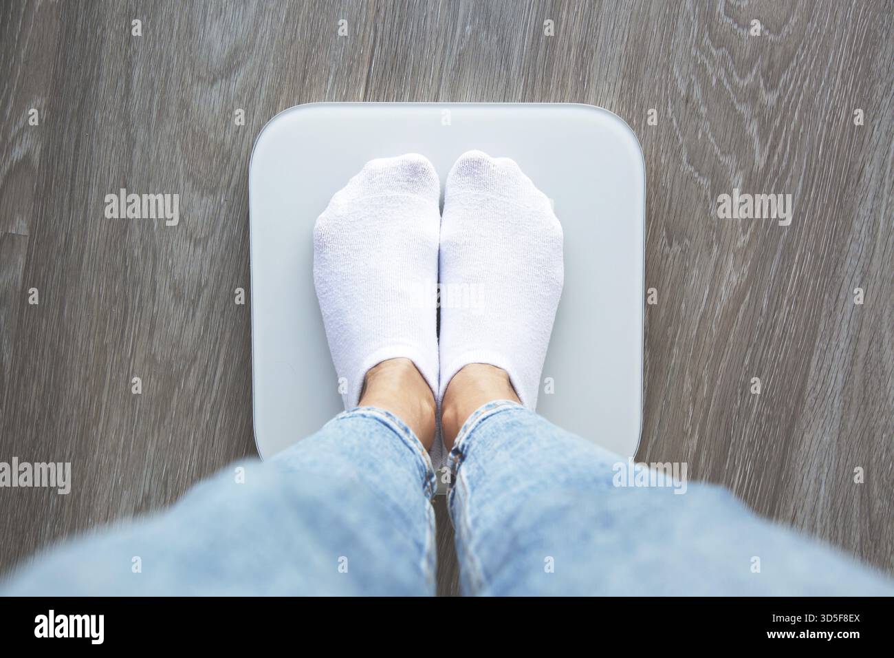 Eine Person mit weißen Socken und Jeans, die auf einer digitalen Waage steht, von oben betrachtet, symbolisiert Gewichtsmessung und Gesundheitsüberwachung Stockfoto