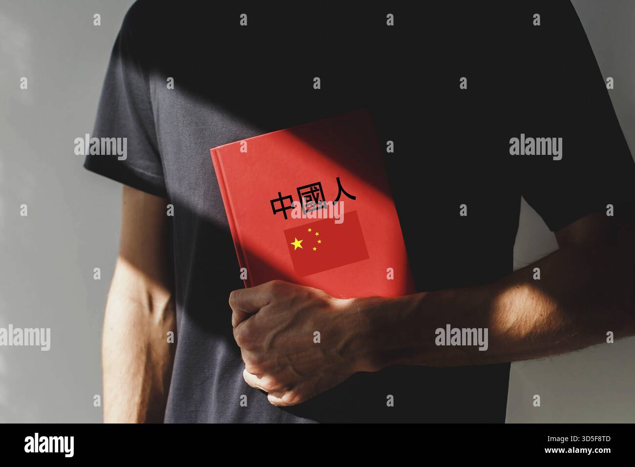 Eine Person, die ein schwarzes T-Shirt trägt, hält ein rotes chinesisches Lehrbuch mit chinesischen Schriftzeichen und einer chinesischen Flagge auf dem Cover und steht im Sonnenlicht. Stockfoto