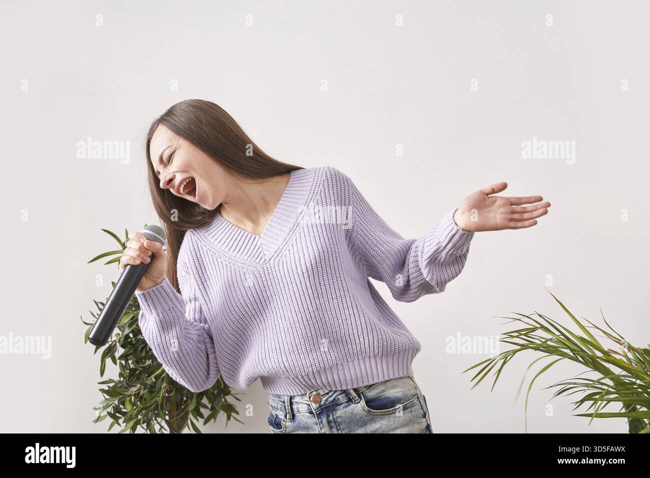 Die junge Frau singt zu Hause. Schwarzes Karaoke-Mikrofon. Die Texte des Hauses tanzen. Lustiges Lied. Unabhängiger Lebensstil zu Hause. Quarantäneaktion sitzend. Lila Dres Stockfoto