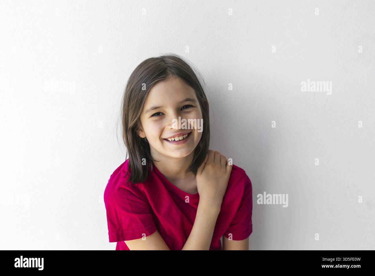 Glückliches Mädchen mit hellem Lächeln und Hand auf Schulter vor weißer Wand, Vorstellung von Kinderfreude und Positivität Stockfoto