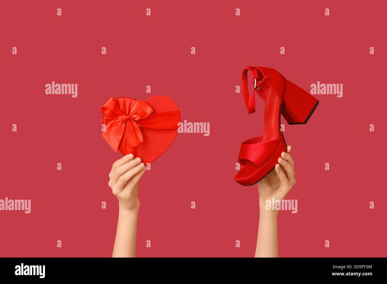Weibliche Hände mit herzförmiger Geschenkbox und Absatzschuh auf rotem Hintergrund. Valentinstag Stockfoto