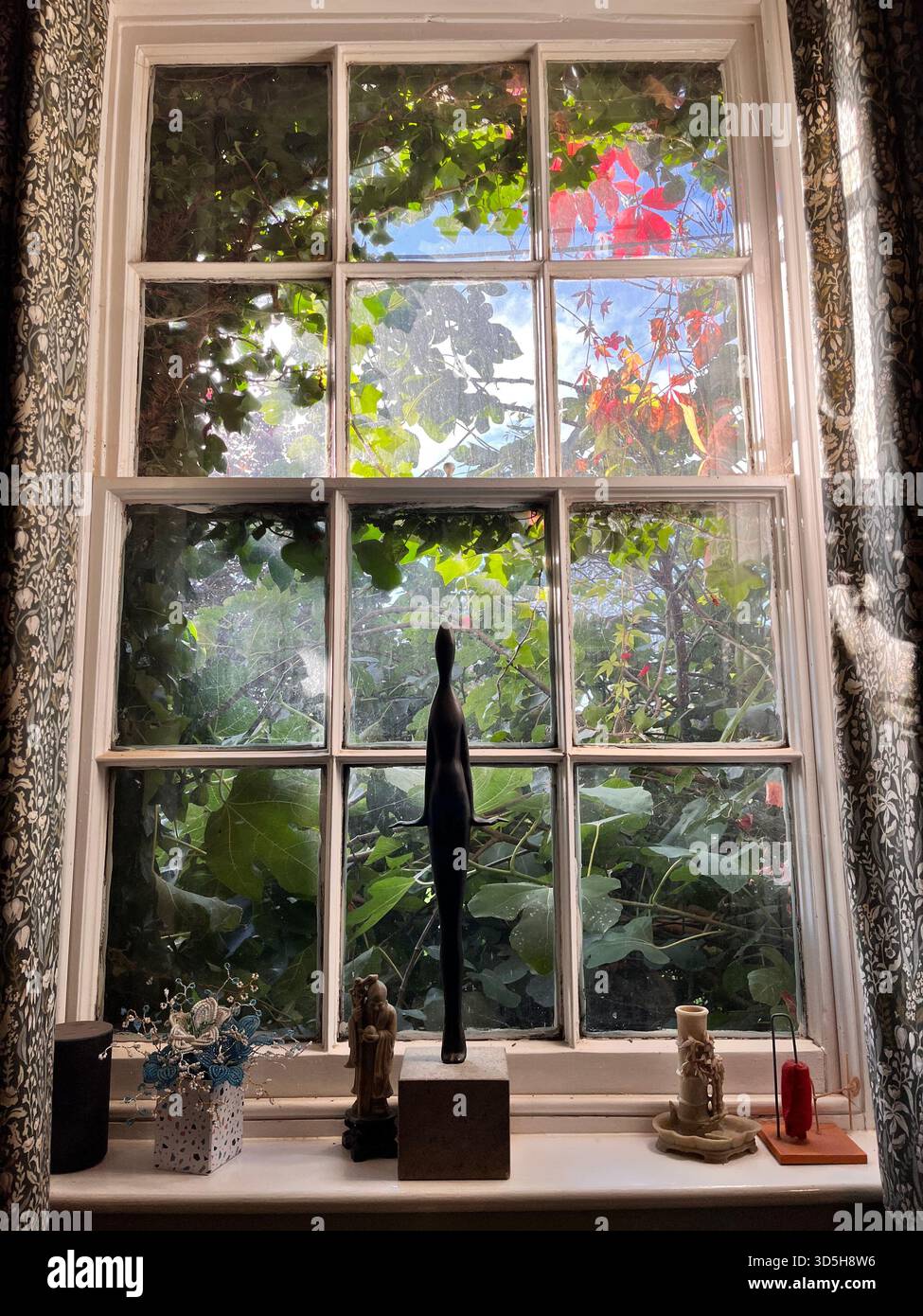 Sonnenlicht strömt durch ein Fenster, das von Pflanzen und dekorativen Gegenständen umgeben ist. Warme, gemütliche Inneneinrichtung mit Gartenblick - Smartphone-aufgenommenes Stockfoto