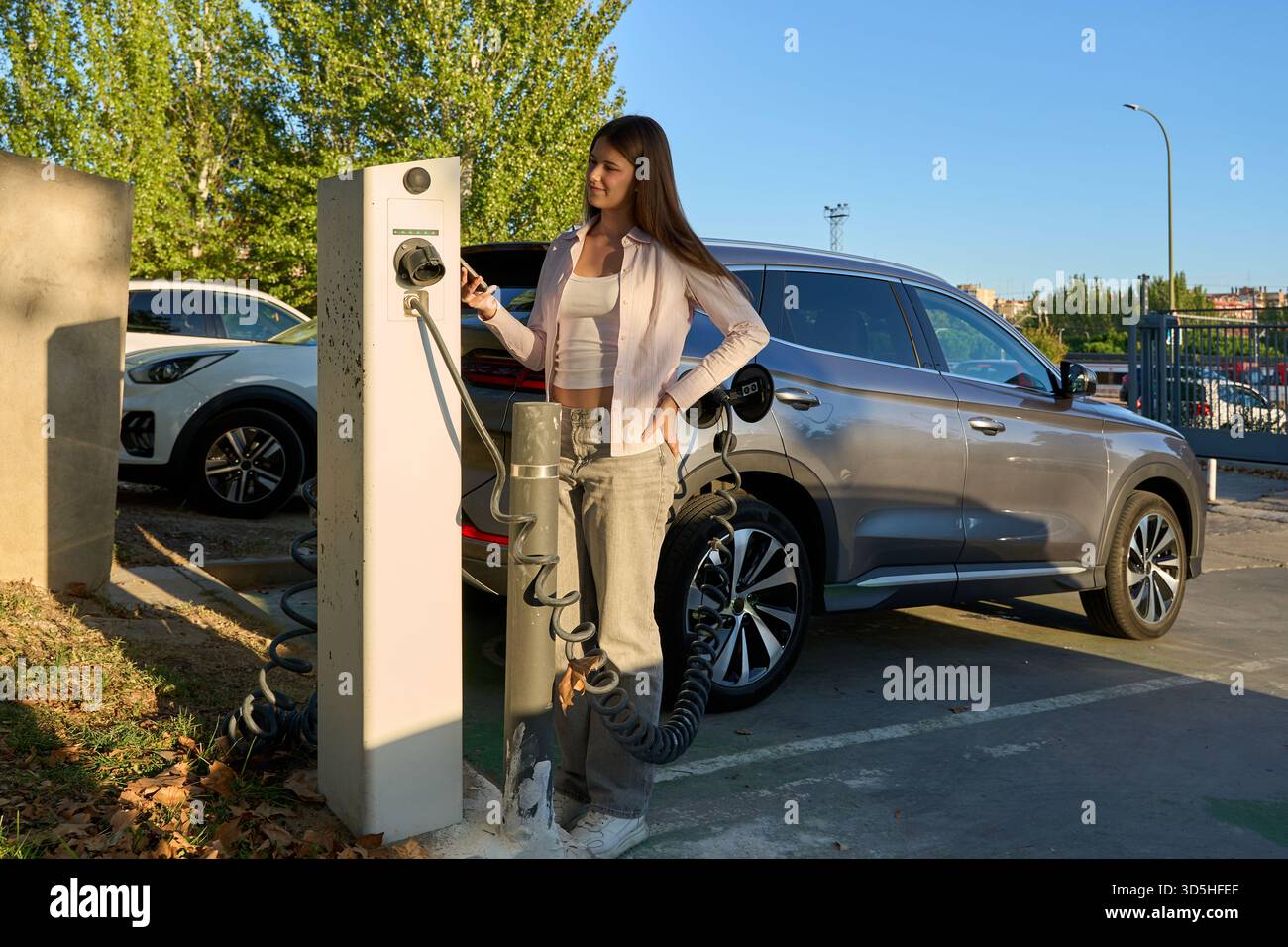 Eine junge Frau, die neben einer EV-Ladestation steht, ihr Telefon benutzt, während ein Elektroauto aufgeladen wird, um ein umweltfreundliches Leben zu demonstrieren Stockfoto