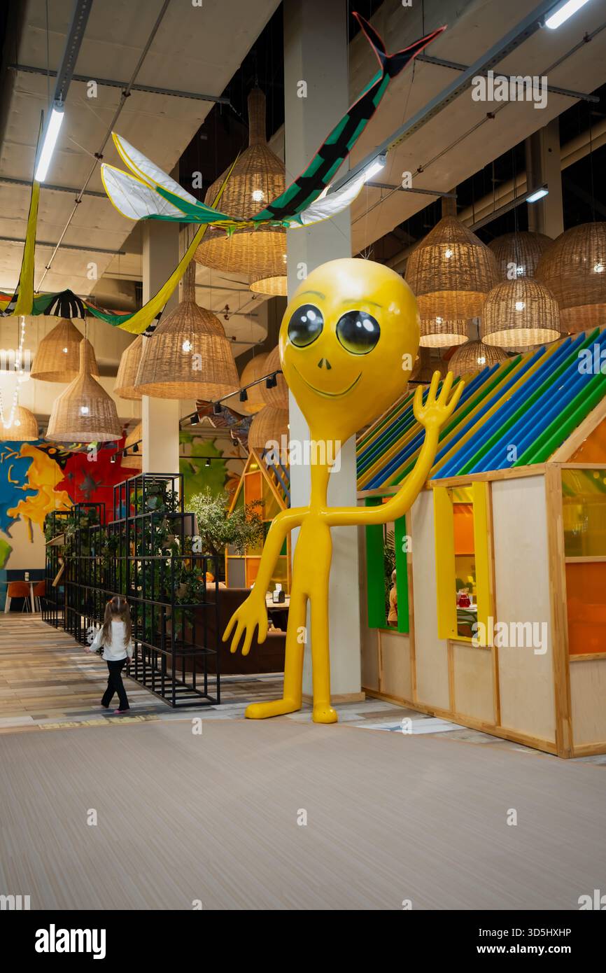 Große, lächelnde Skulptur neben einem Kinderspielhaus in einem modernen, von warmer Beleuchtung und Spielfreude umgebenen Indoor-Aktivitätenzentrum Stockfoto