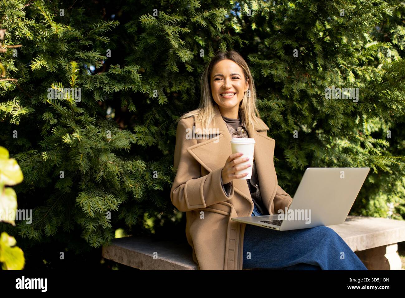 Eine Frau sitzt draußen und genießt einen Kaffee, während sie an ihrem Laptop arbeitet. Umgeben von lebendigem Grün, lächelt sie und spiegelt ein entspanntes, produktives atmos wider Stockfoto