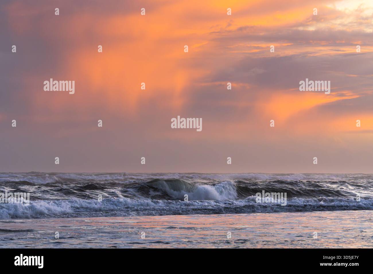 Meereswellen krachen bei Sonnenuntergang unter leuchtenden orange und violetten Wolken Stockfoto