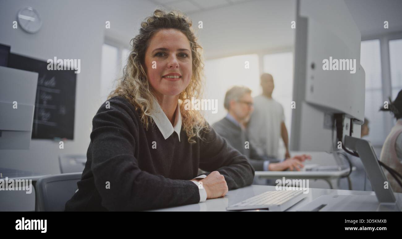 Lockhaarige Frau besucht den Coding Workshop, schaut auf den Bildschirm und lächelt warm. Konzept kreatives Lernen, positive Bildungsatmosphäre, Sand Digital Empowerment im modernen Klassenzimmer. Hochformat. Stockfoto