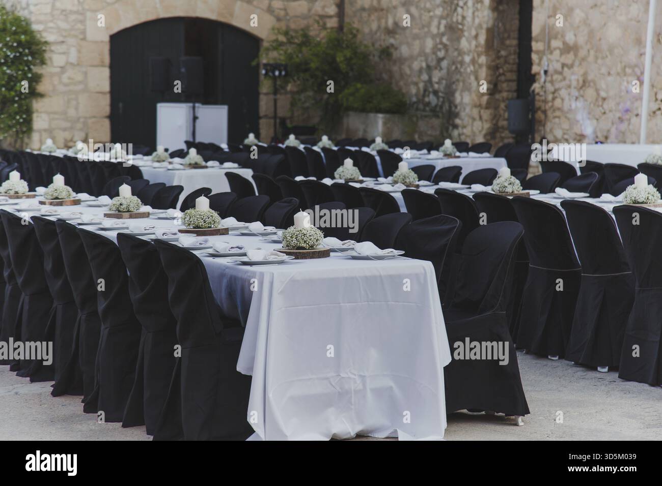 Elegante Banketteinrichtung im Freien mit langen Tischen, die mit weißen Tischdecken bedeckt sind, schwarzen Stühlen und geblümten Mittelstücken vor rustikaler Steinkulisse Stockfoto