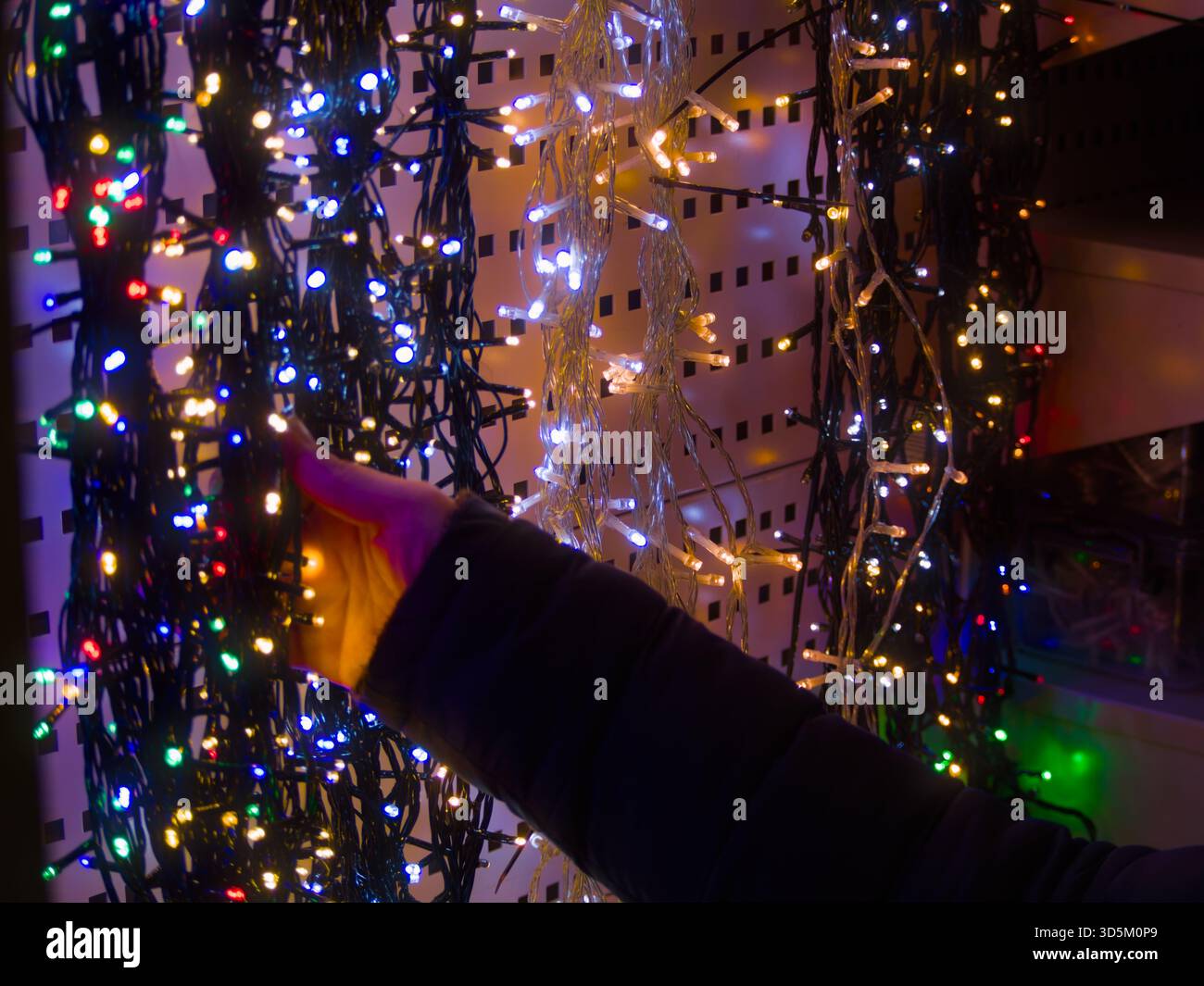 Weihnachtslichter im Geschäft mit der Hand berühren – die Hand Einer Person reicht aus, um einen Vorhang aus farbenfrohen, unscharfen Weihnachtslichtern zu berühren Stockfoto