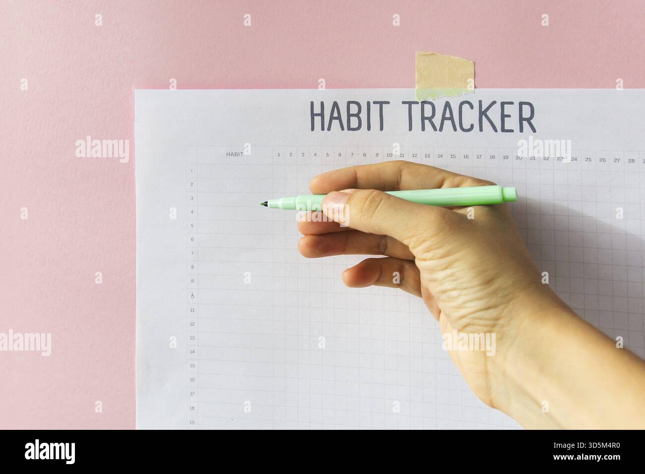 Hand mit einem Stift auf dem Hintergrund eines Habit Trackers, der an einer pinkfarbenen Wand hängt, Konzept der Motivation und Selbstentwicklung Stockfoto