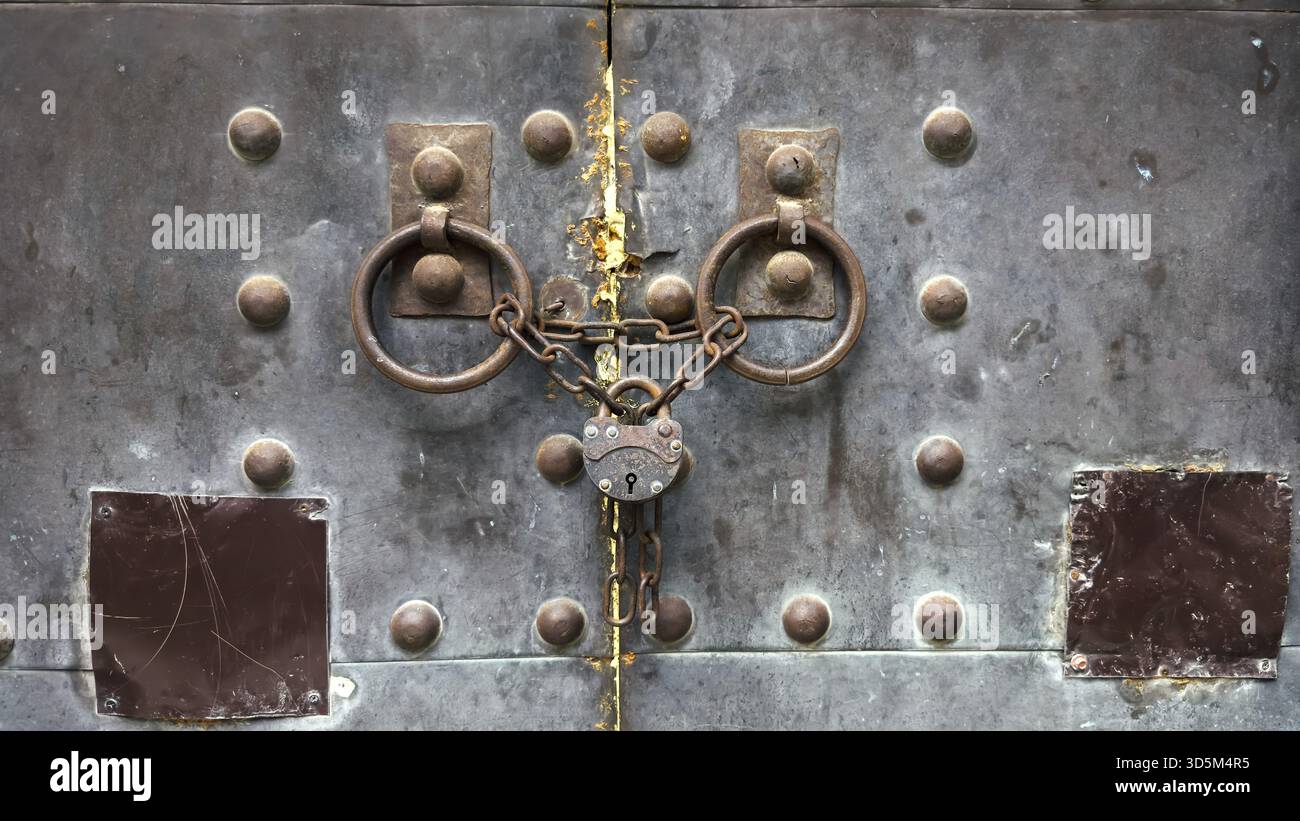 Eine Nahaufnahme einer alten Metalltür, die mit rostigen Ketten und einem alten Vorhängeschloss gesichert ist, mit schweren Eisenringen und verwitterten Texturen mit Zeichen des Alters A Stockfoto