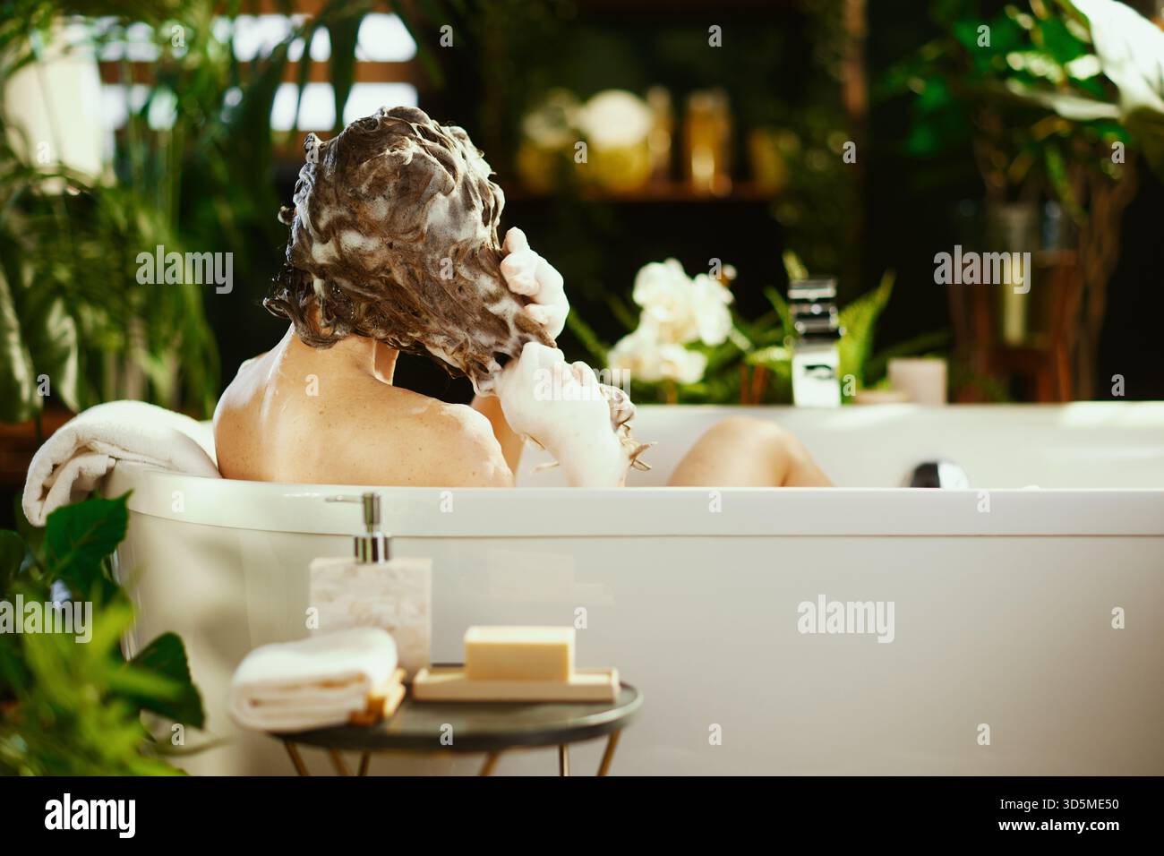 Person wäscht Haare mit Shampoo in einer weißen Badewanne, umgeben von üppig grünen Pflanzen. Weiße Handtücher und Badartikel in der Nähe schaffen einen ruhigen, Spa-ähnlichen geldautomaten Stockfoto