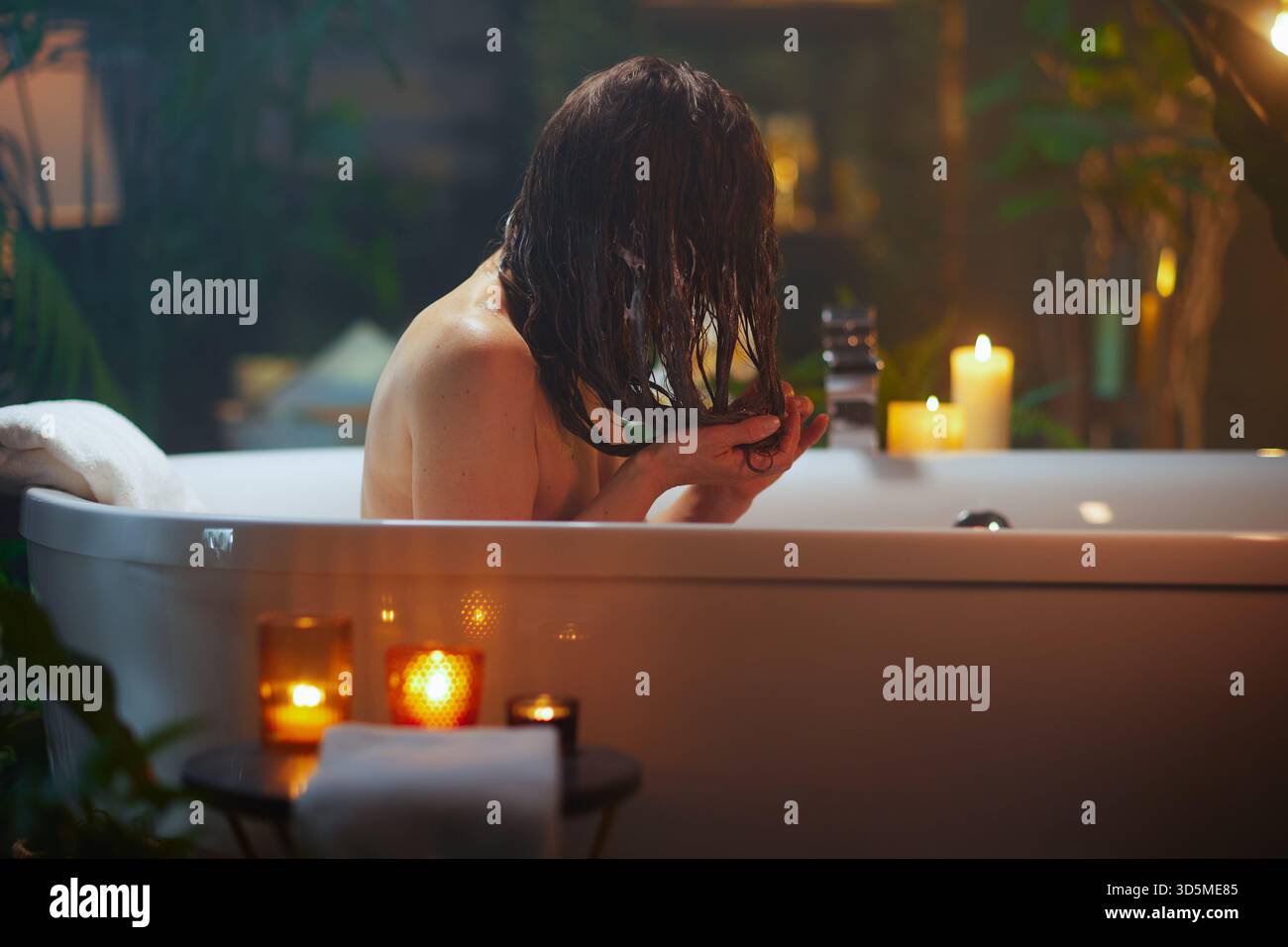 Eine Person, von hinten gesehen, mit langen, nassen, dunklen Haaren, hält es wie beim Spülen oder Auftragen der Haarmaske in einer Badewanne. Sanfte Beleuchtung durch Kerzen und umgeben von B Stockfoto