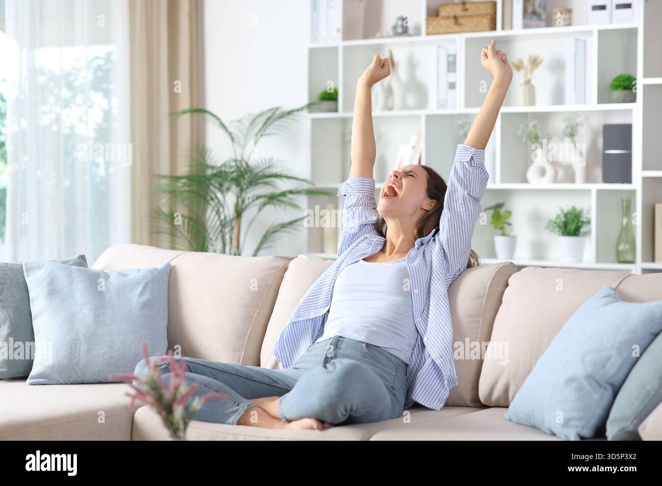 Aufgeregte Frau in blauen Armen, die Erfolg feiert und zu Hause auf einer Couch sitzt Stockfoto