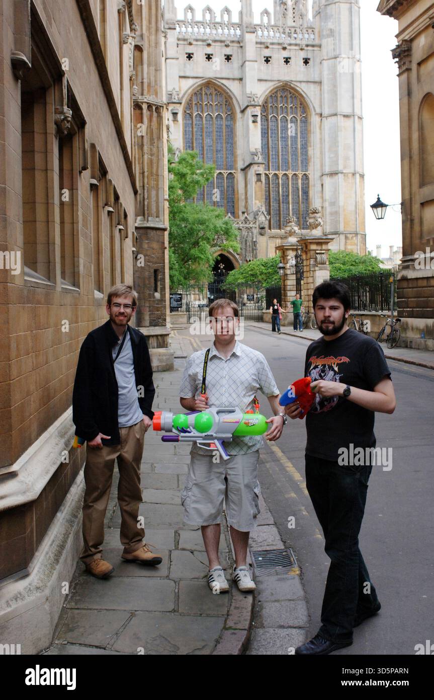 Junge Männer mit farbenfrohen Wasserpistolen auf einer historischen Straße in Cambridge während der Maiball-Feierlichkeiten, die in der Nähe der berühmten College-Architektur stehen Stockfoto